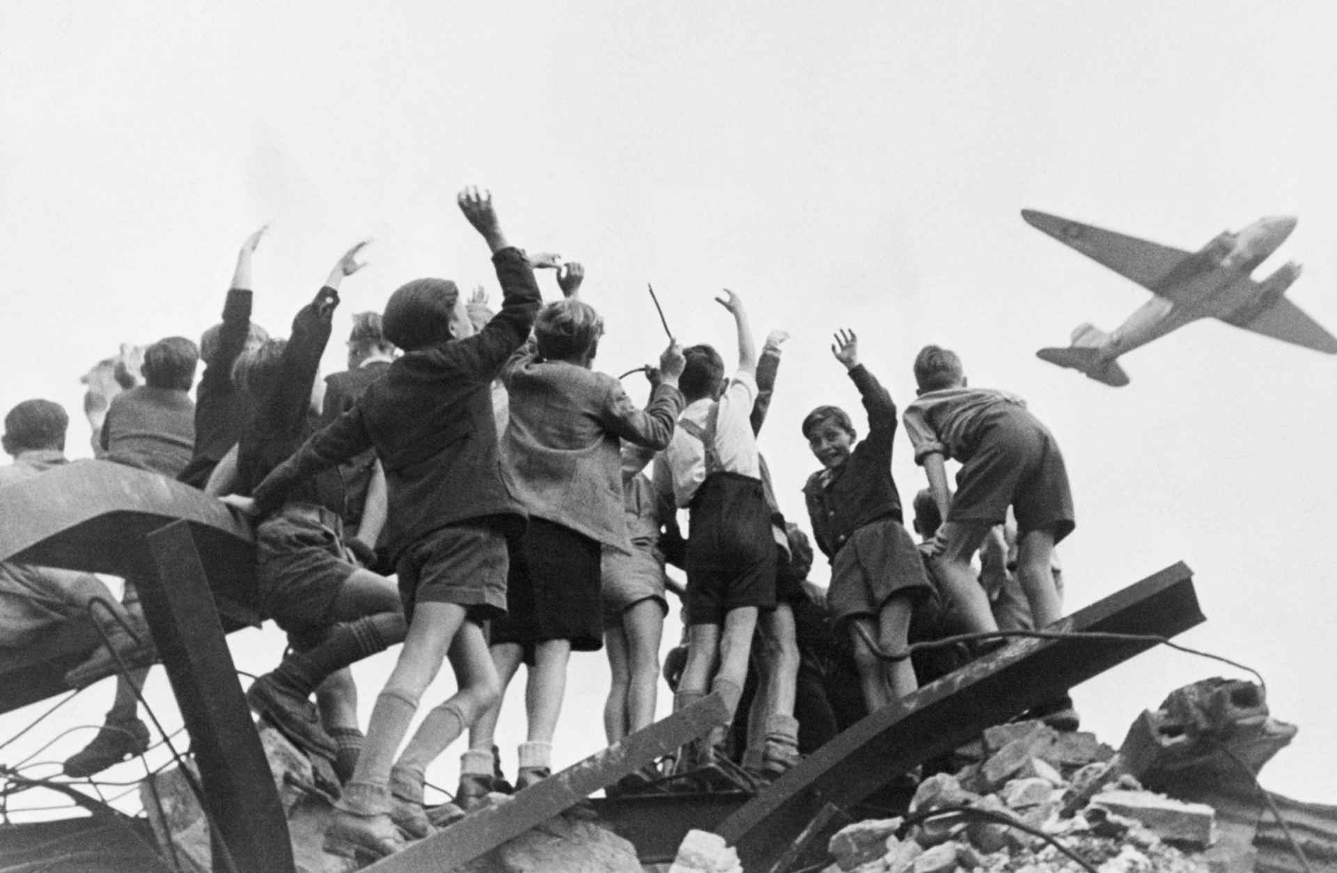 Berlin Airlift - A group of German children stand atop building rubble, cheering a U.S. cargo airplane as it flies over a western section of Berlin in January 1948.