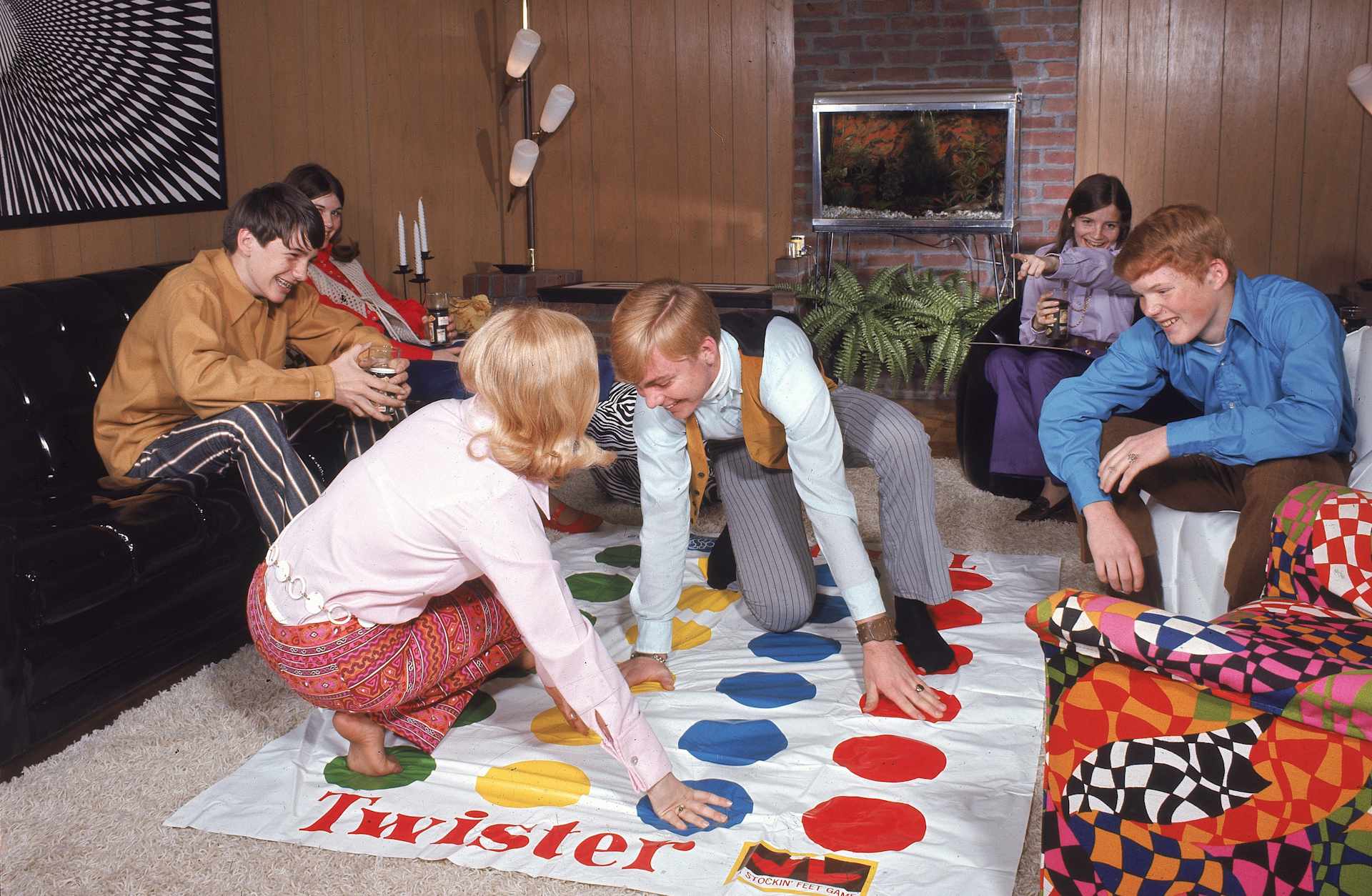 A boy and girl play the game Twister, as other young people watch in a paneled living room, c. 1968.