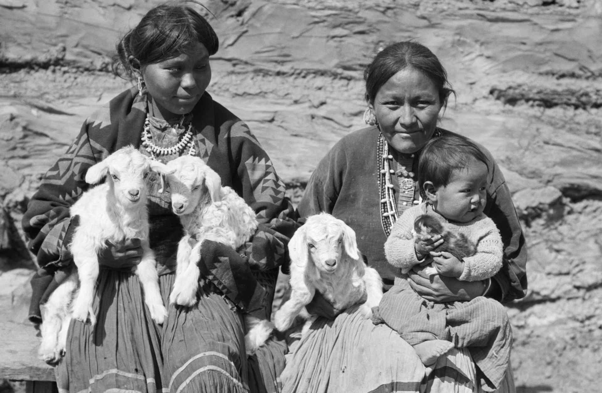 Two Navajo women, pictured with a baby and three small lambs, c. 1930s.