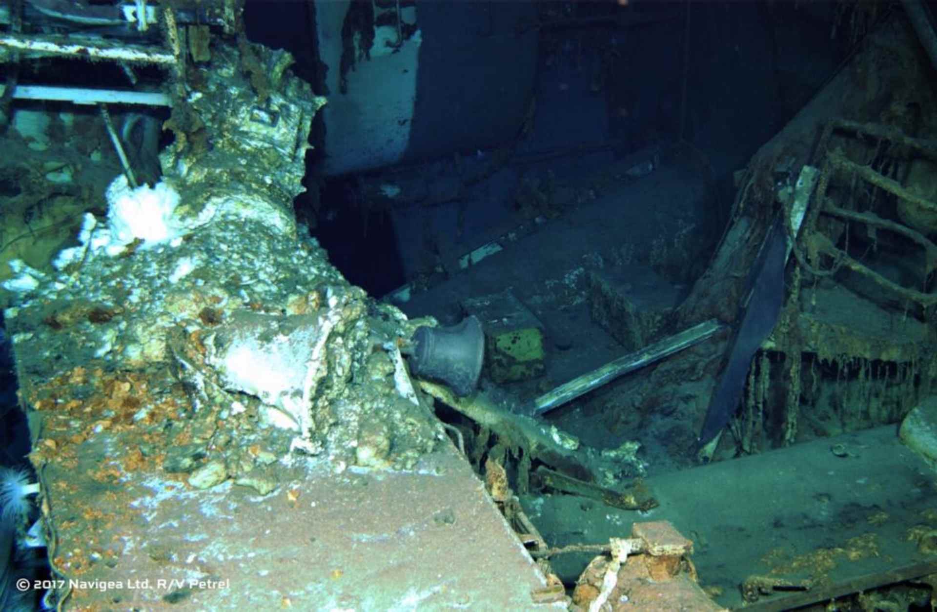 The image depicts a submerged shipwreck, with the foreground showing a heavily damaged and overgrown structure, while the background is shrouded in darkness, suggesting a deep underwater environment.