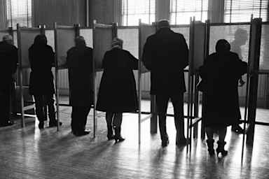Circa 1970s: Men and women standing side by side in private voting booths, filling out election ballot papers.