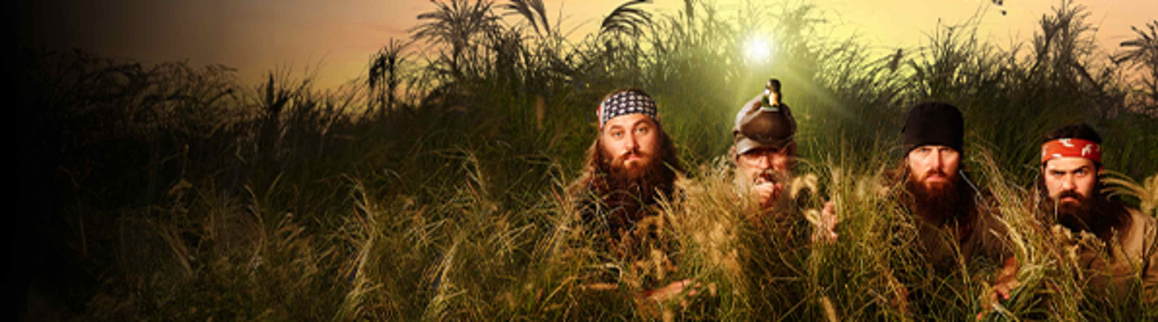 Two men with long beards and traditional headwear stand in a field of tall grass, with a warm, hazy background.