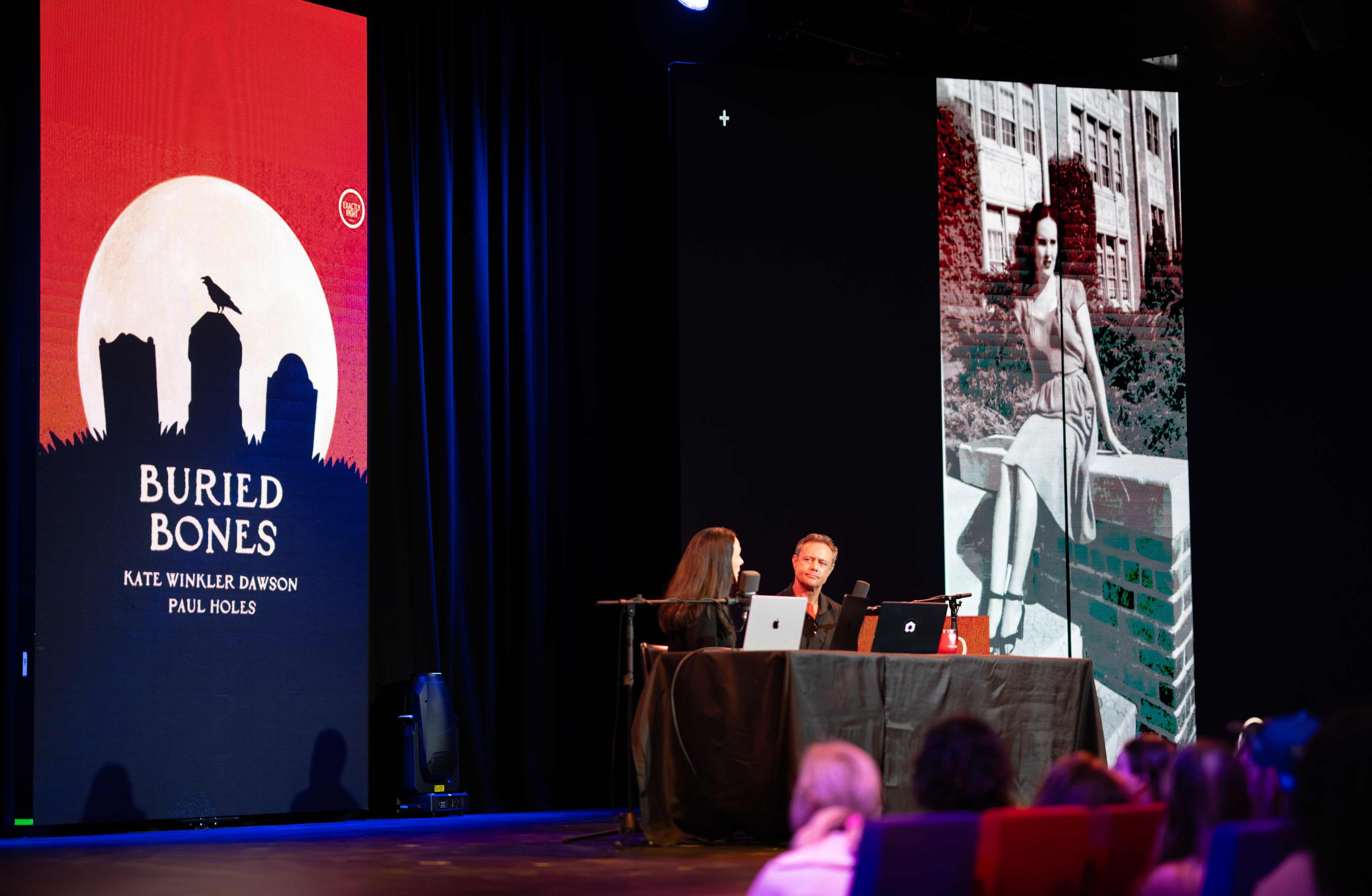 The image shows a stage with a large red banner displaying the title "Buried Bones" and two people, likely speakers, seated at a table in the foreground.