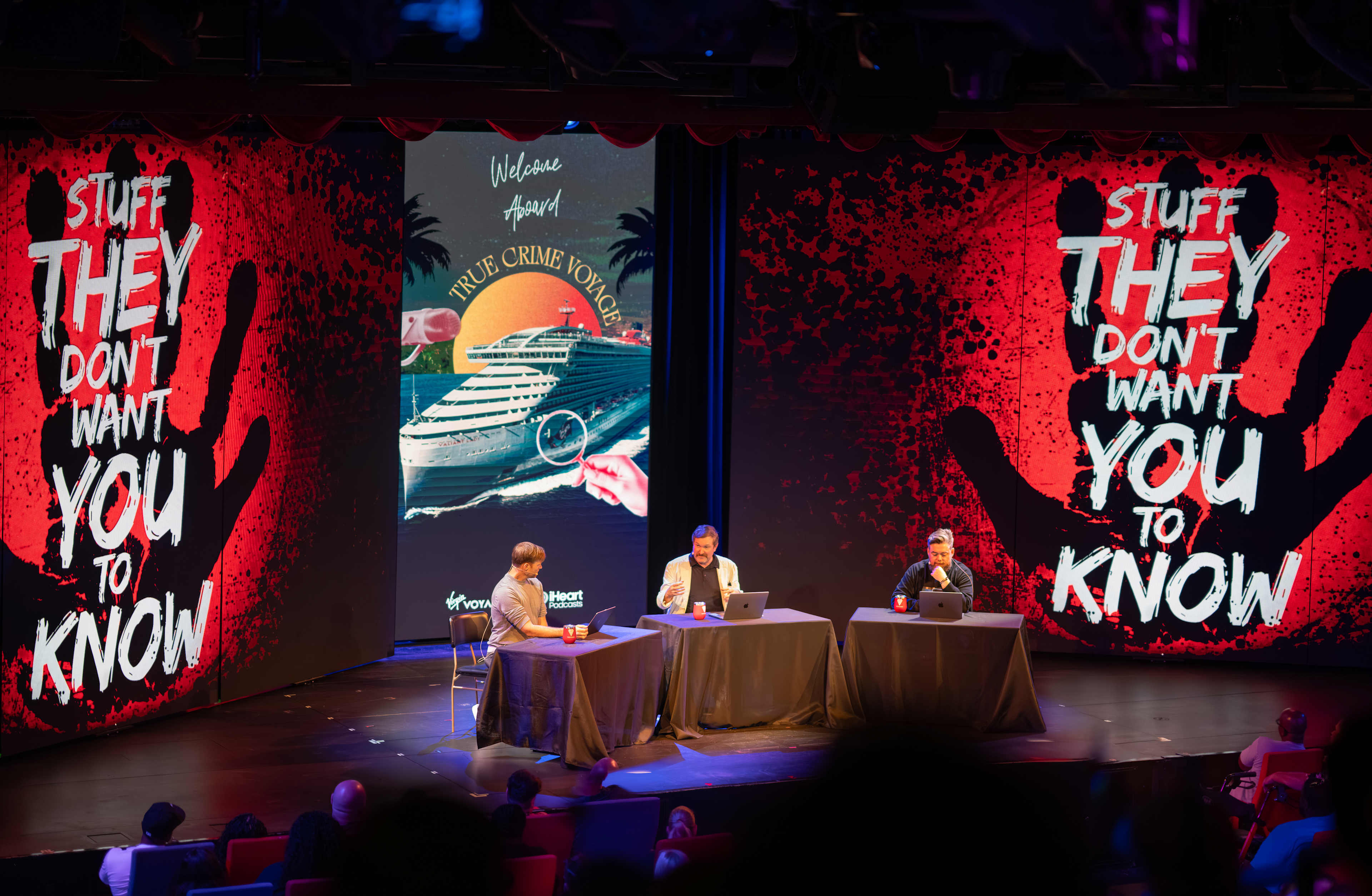 The image shows a stage with three people sitting at a table, surrounded by large red and black banners with text that reads "Stuff they don't want you to know" and "Stuff they don't want you to know".
