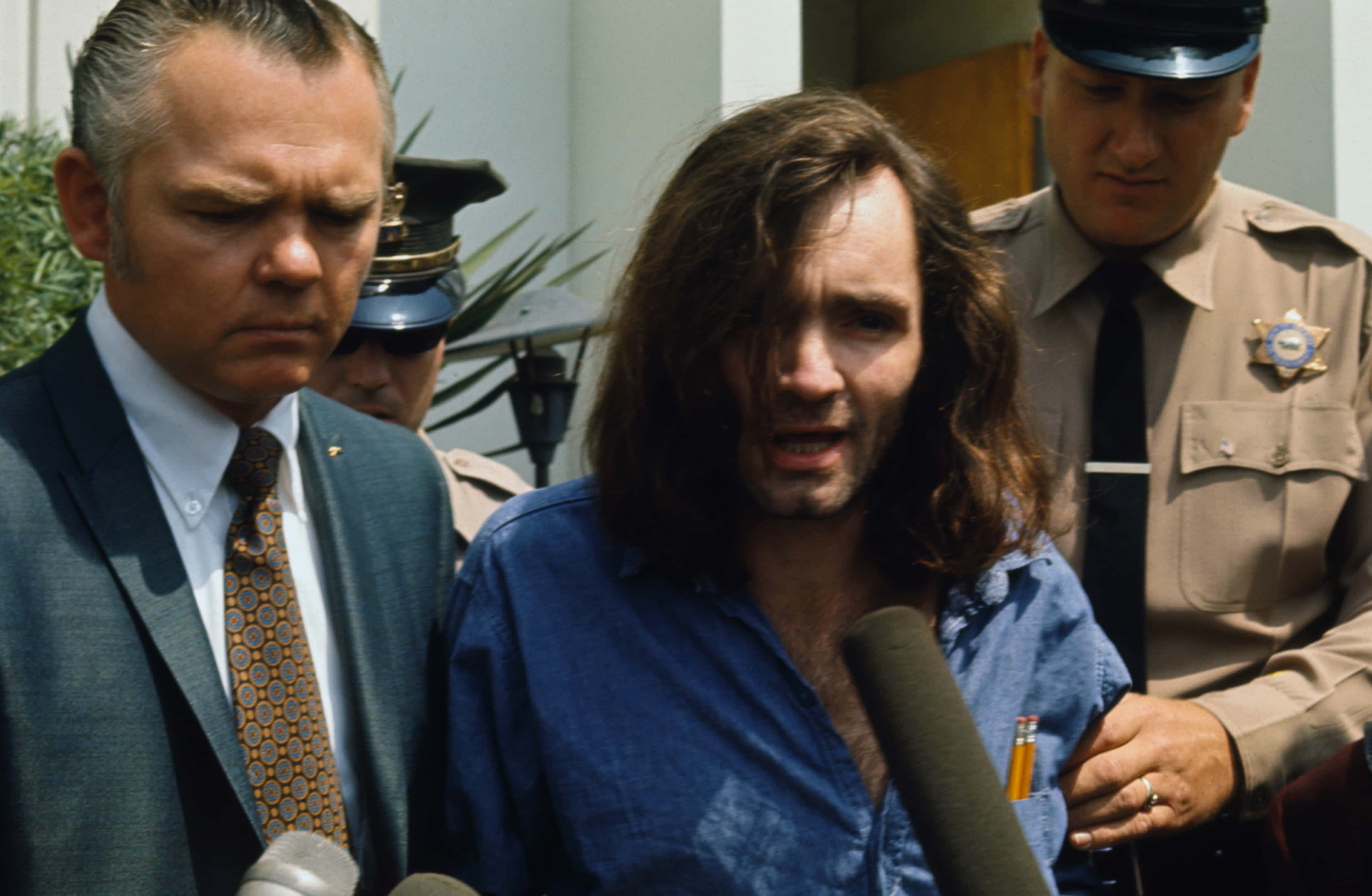 The image shows a young woman being escorted by law enforcement officers, with a serious expression on her face.