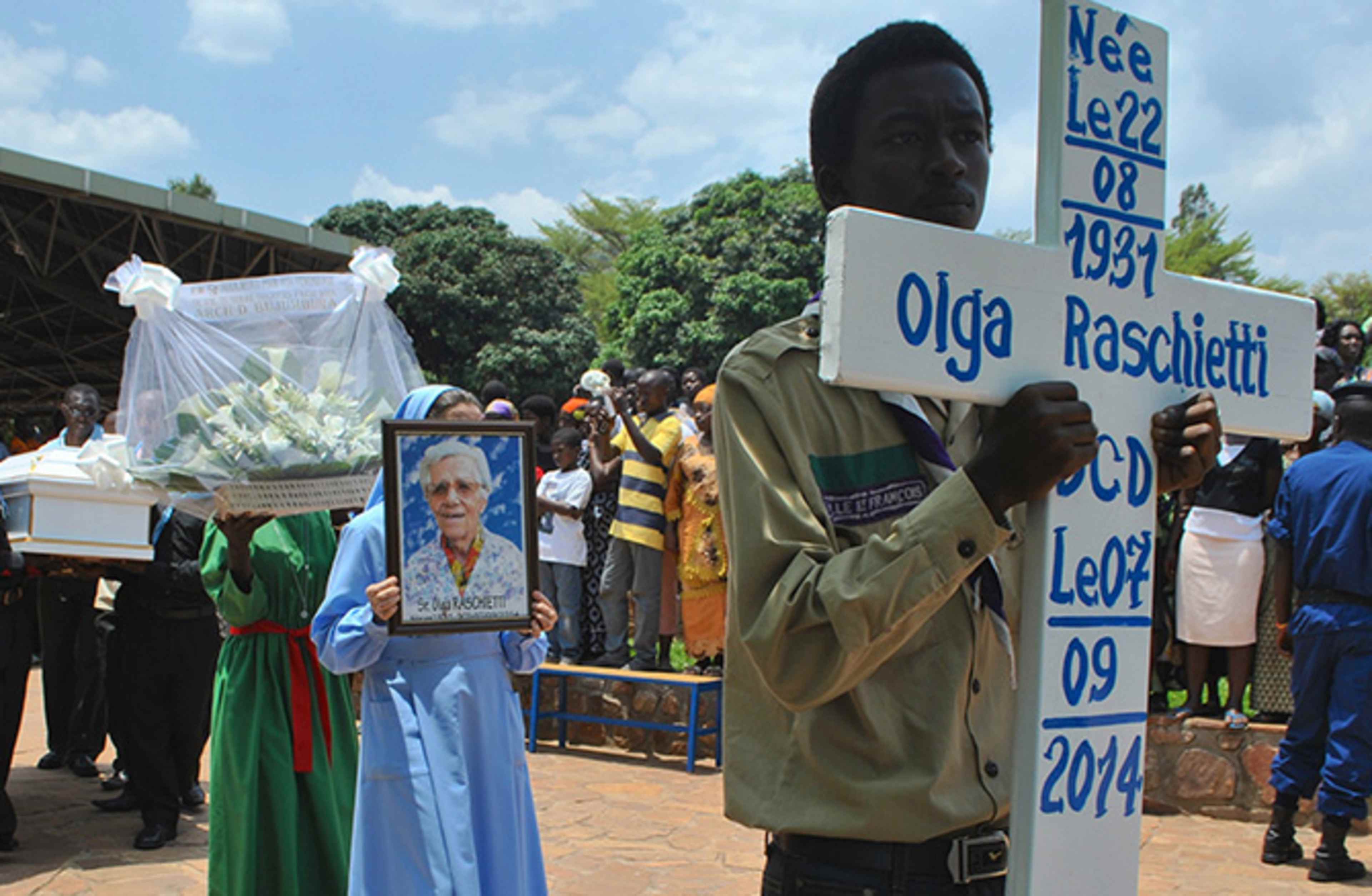 A crowd of people gathered around a memorial display, with a person holding a framed photograph and a sign bearing the name "Olga Raschetti".