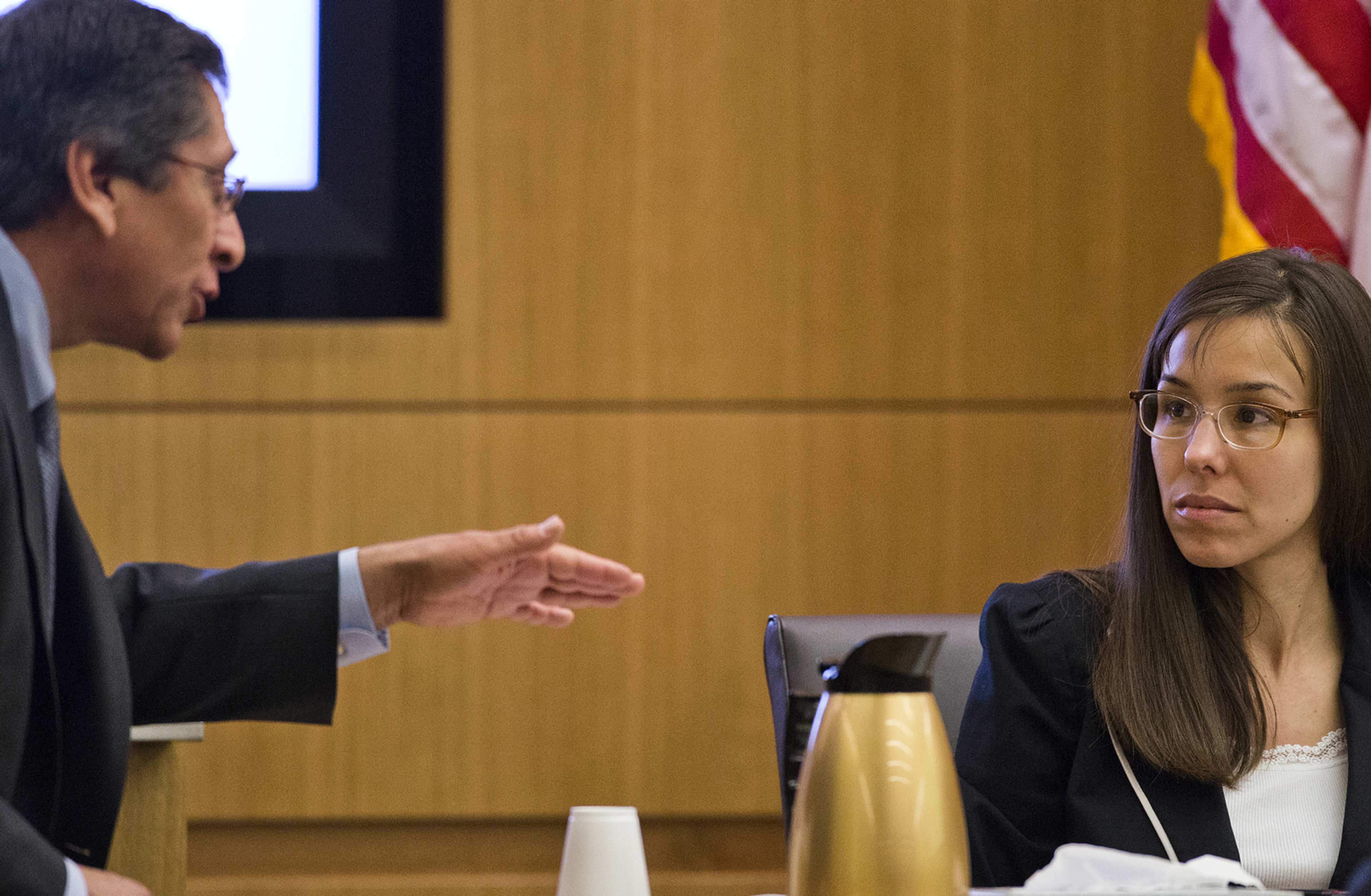 Two individuals, a man and a woman, are engaged in a conversation in what appears to be a courtroom setting, with an American flag visible in the background.