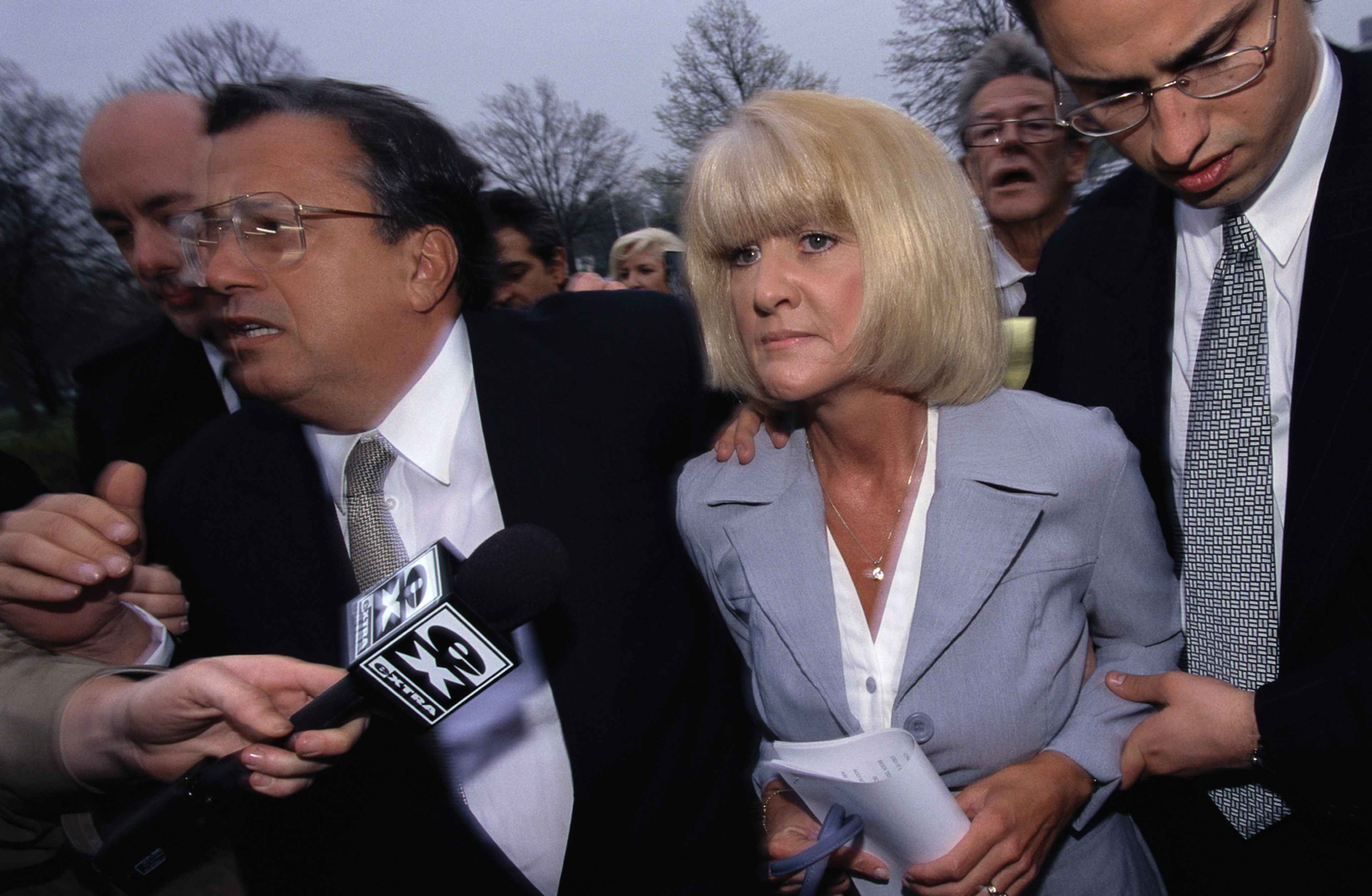 The image shows a group of people, including a woman with blonde hair, walking together in what appears to be a formal setting, with trees and buildings visible in the background.