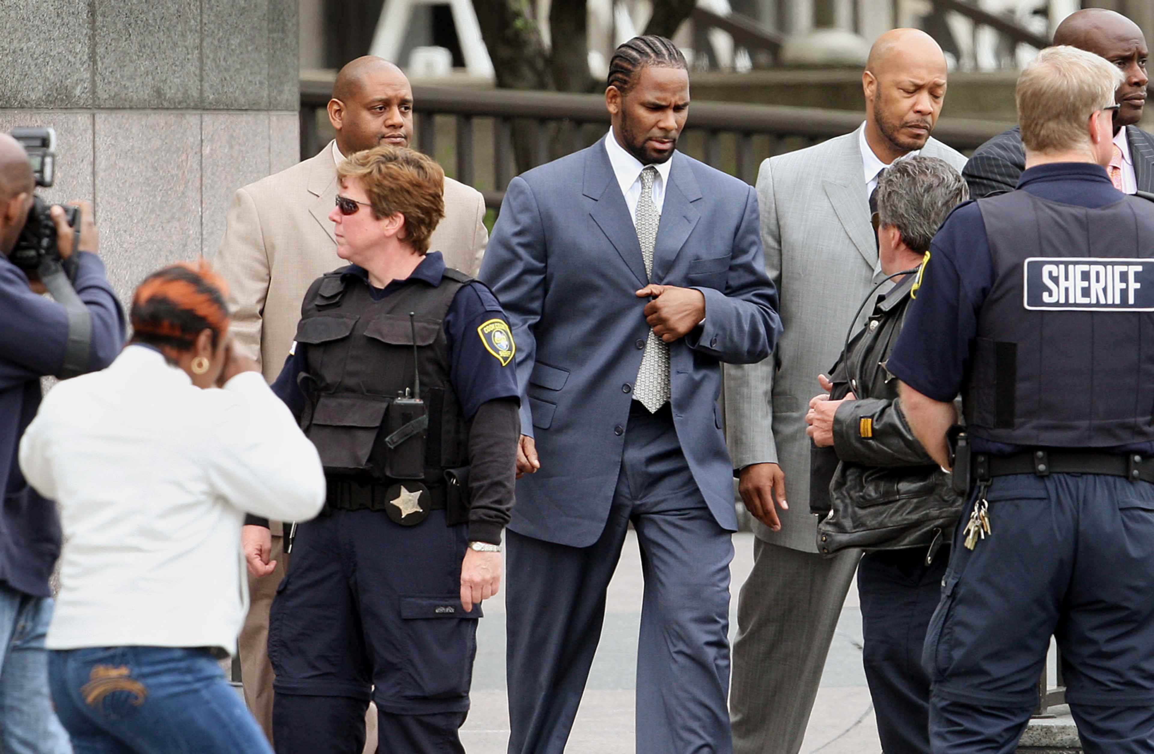 The image shows several individuals, including law enforcement officers and a man in a suit, gathered outside what appears to be a courthouse or government building.