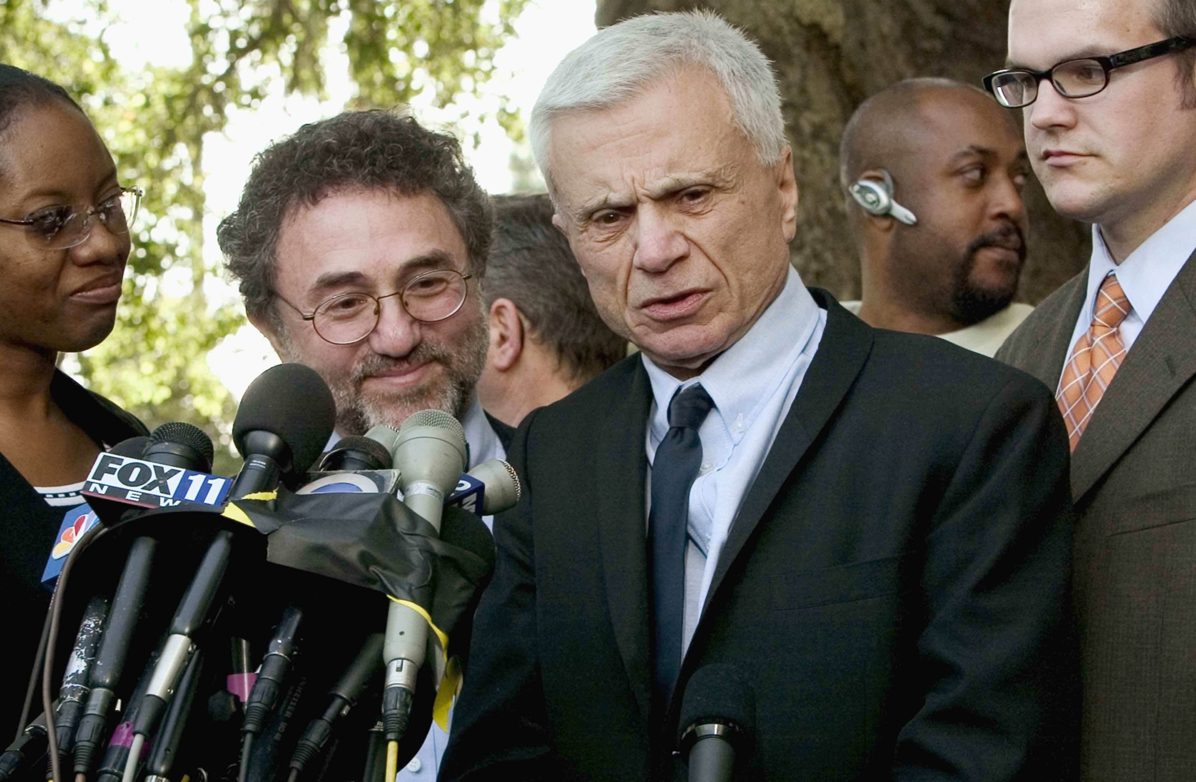 The image shows a group of individuals, including several men in suits, standing together and speaking into microphones, with a backdrop of trees and a building in the background.