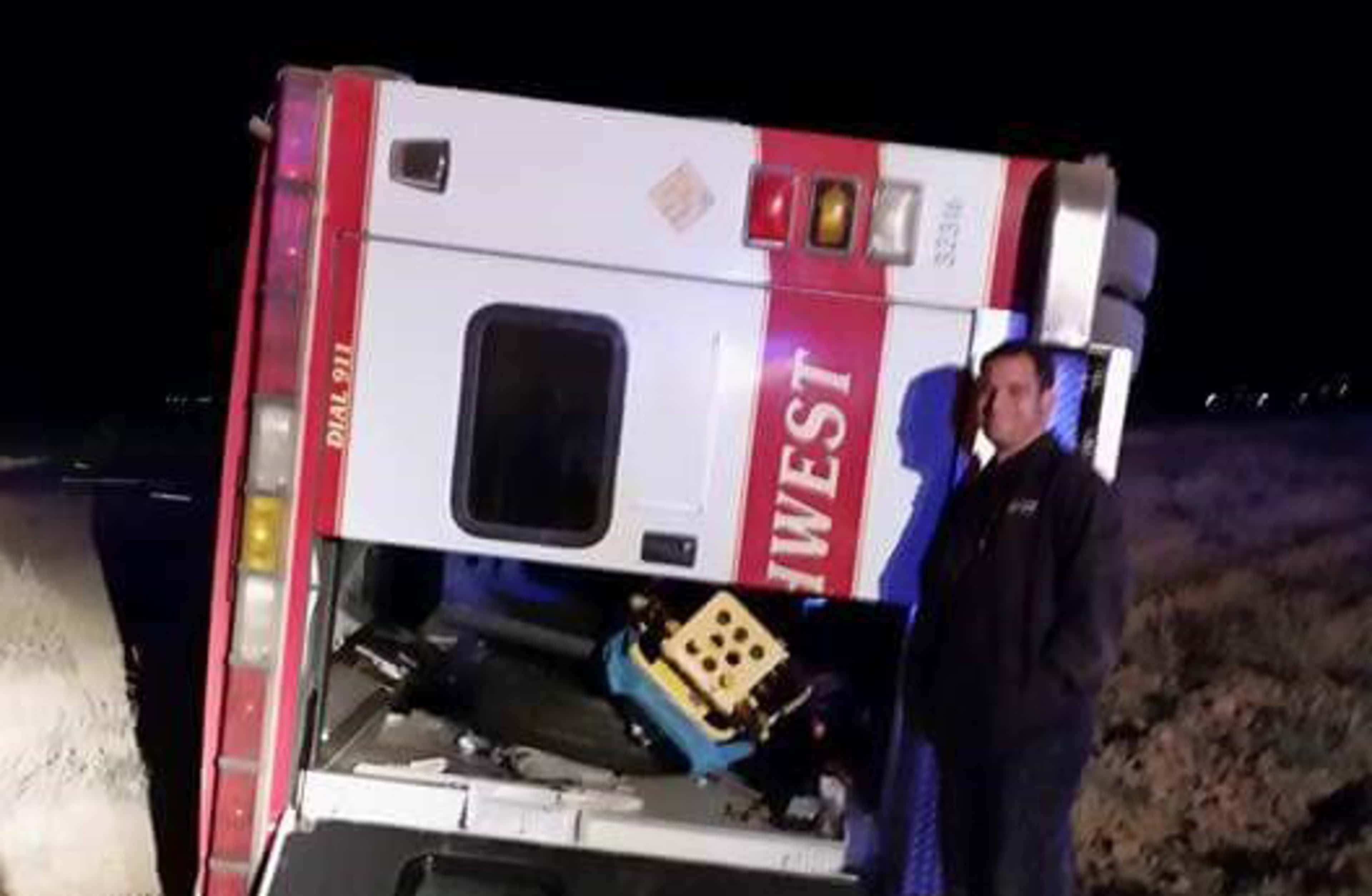 A white emergency medical services vehicle is parked on a dark, snowy road, with a person standing next to it.