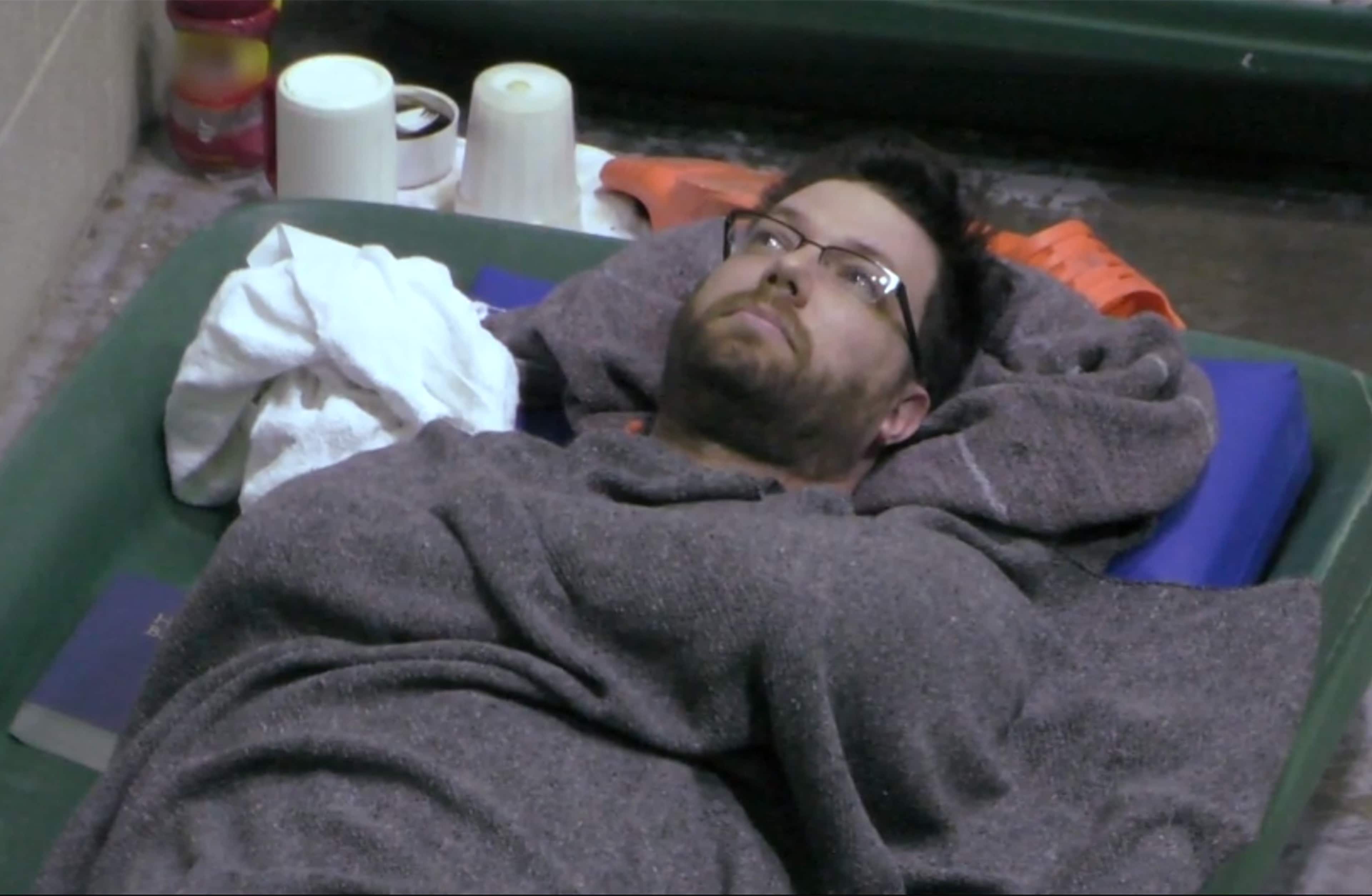 A person with a beard and glasses is lying on a couch or bed, surrounded by various household items in the background.