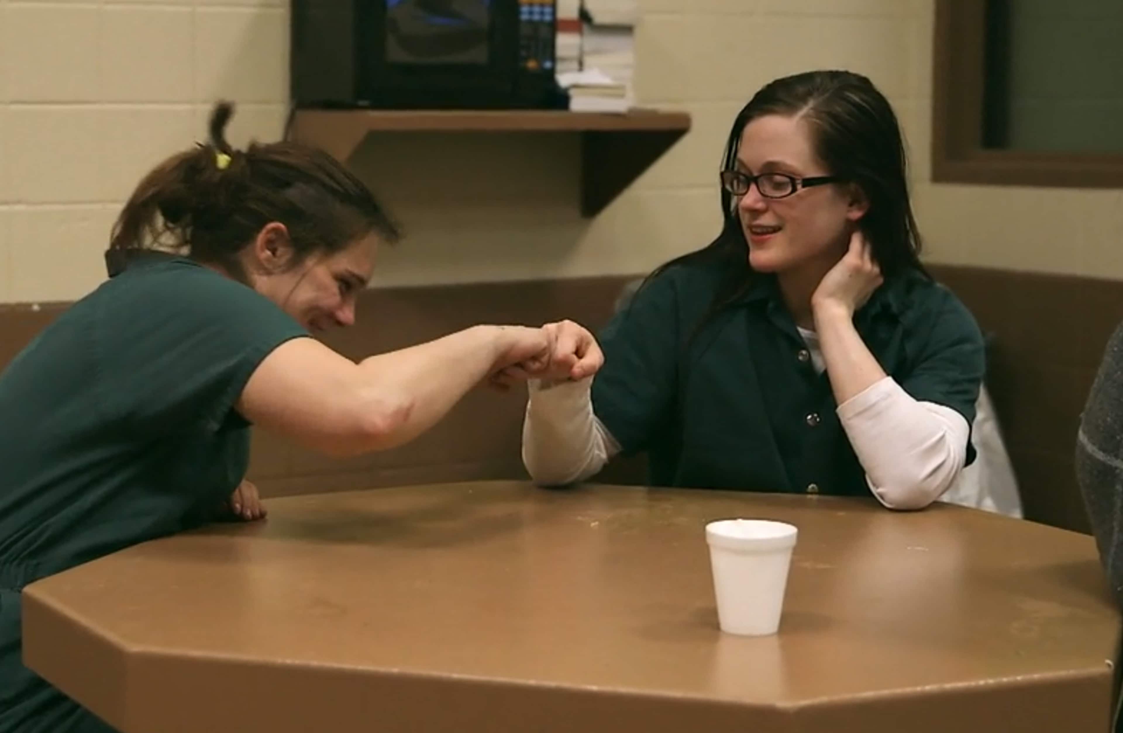 Two individuals, a man and a woman, are engaged in a conversation at a table in what appears to be a casual dining setting, with shelves and other furnishings visible in the background.
