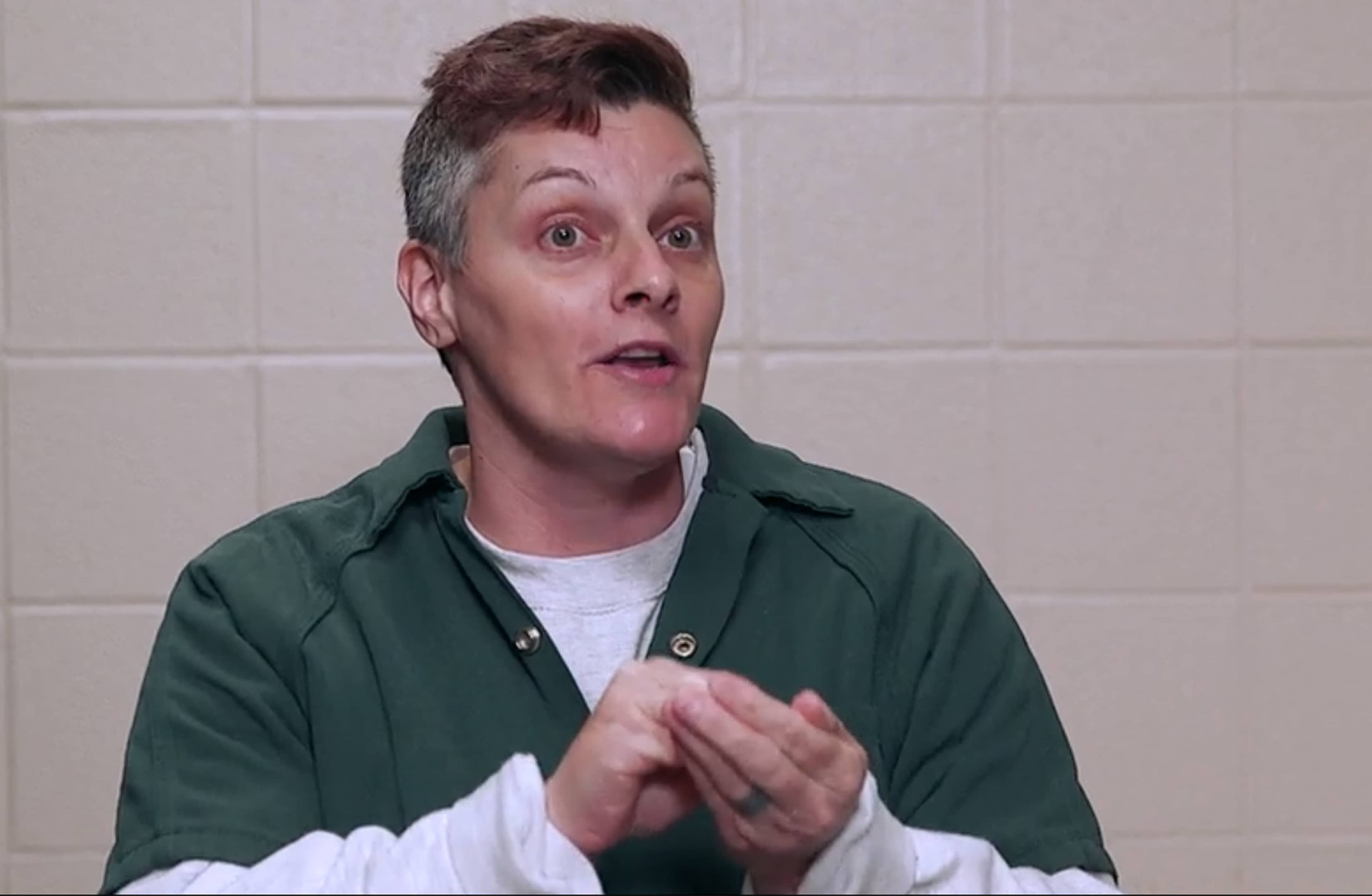 A person with a surprised expression, wearing a green jacket, stands in front of a tiled wall background.