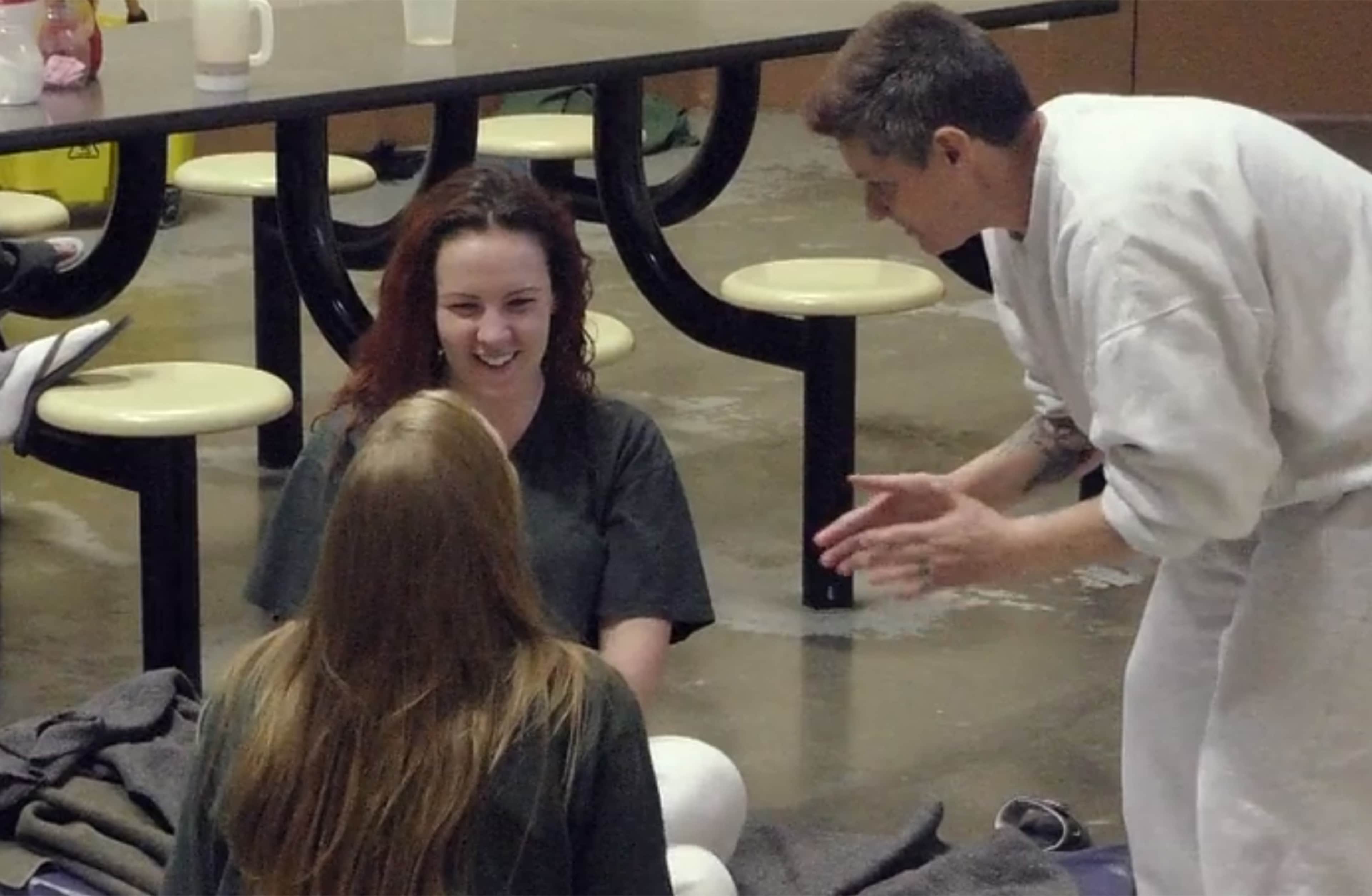 The image shows a woman with long brown hair sitting at a table, smiling and interacting with a man in a white coat, likely a medical professional, who is standing next to her.