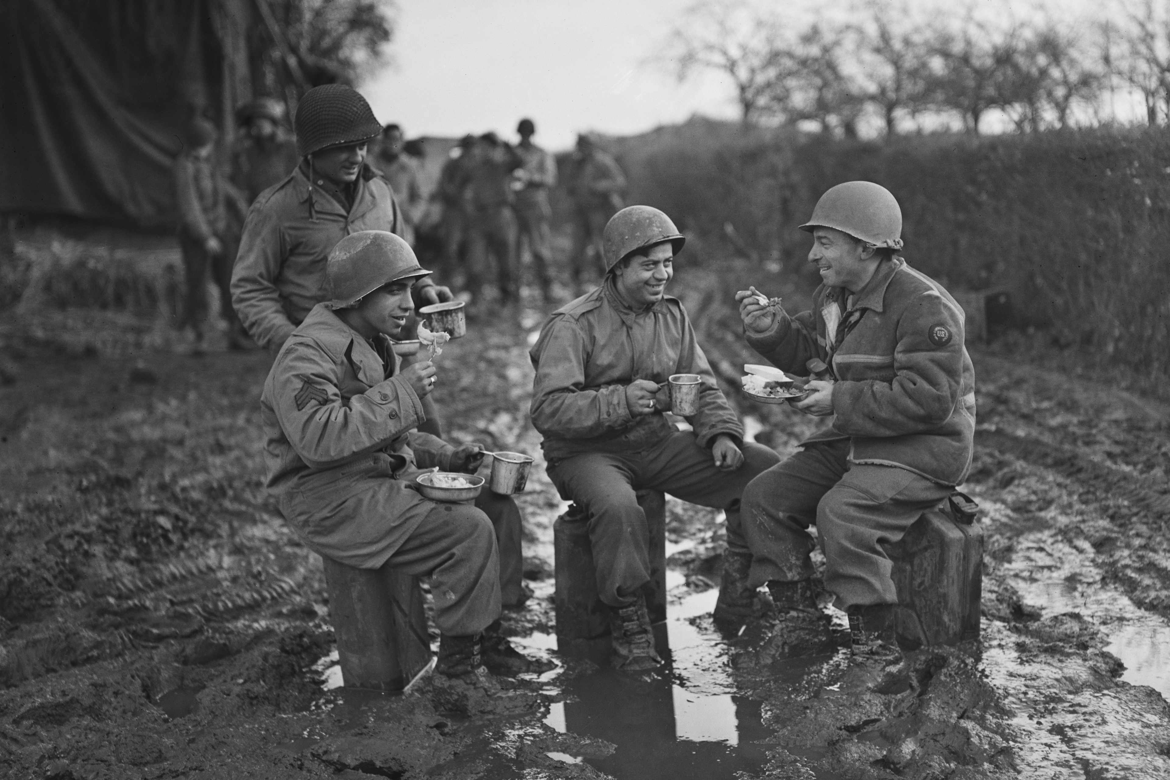 Three US soldiers, one standing and two sitting, smile ate a combat photographer, who is also seated. The three men sitting eat a meal during a World War II campaign in Normandy, France.