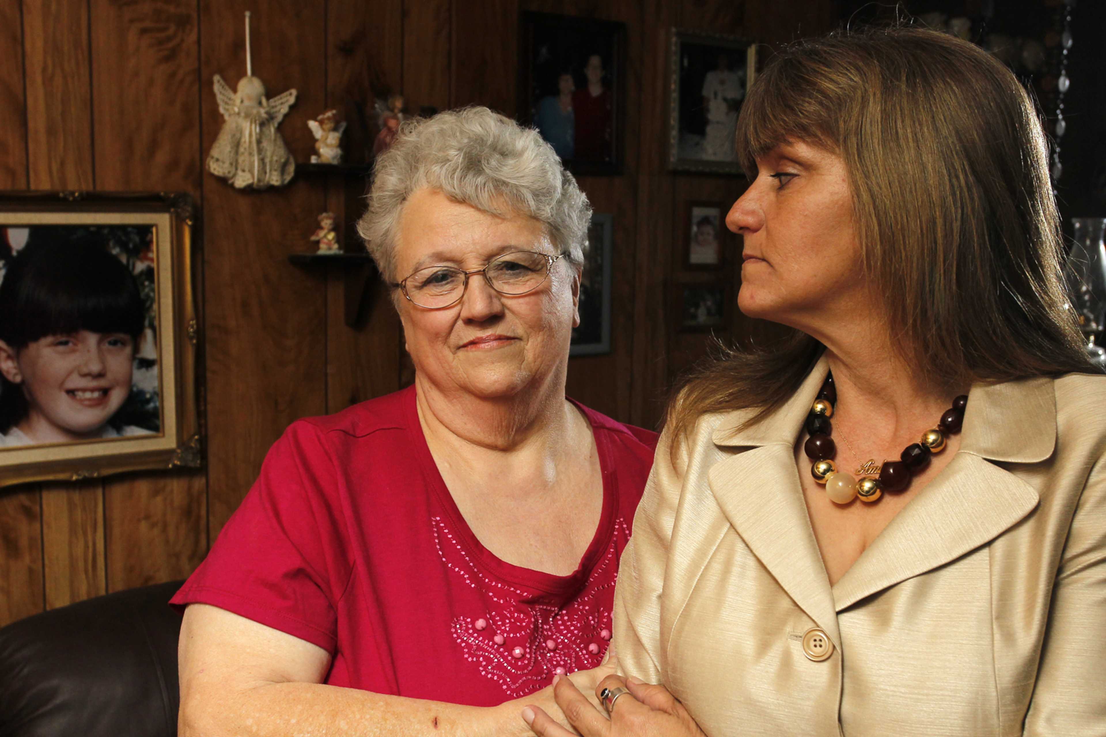 The image shows an older woman with gray hair wearing a red shirt and a younger woman with long brown hair wearing a beige coat, standing together in what appears to be a home setting with framed photographs on the wall behind them.