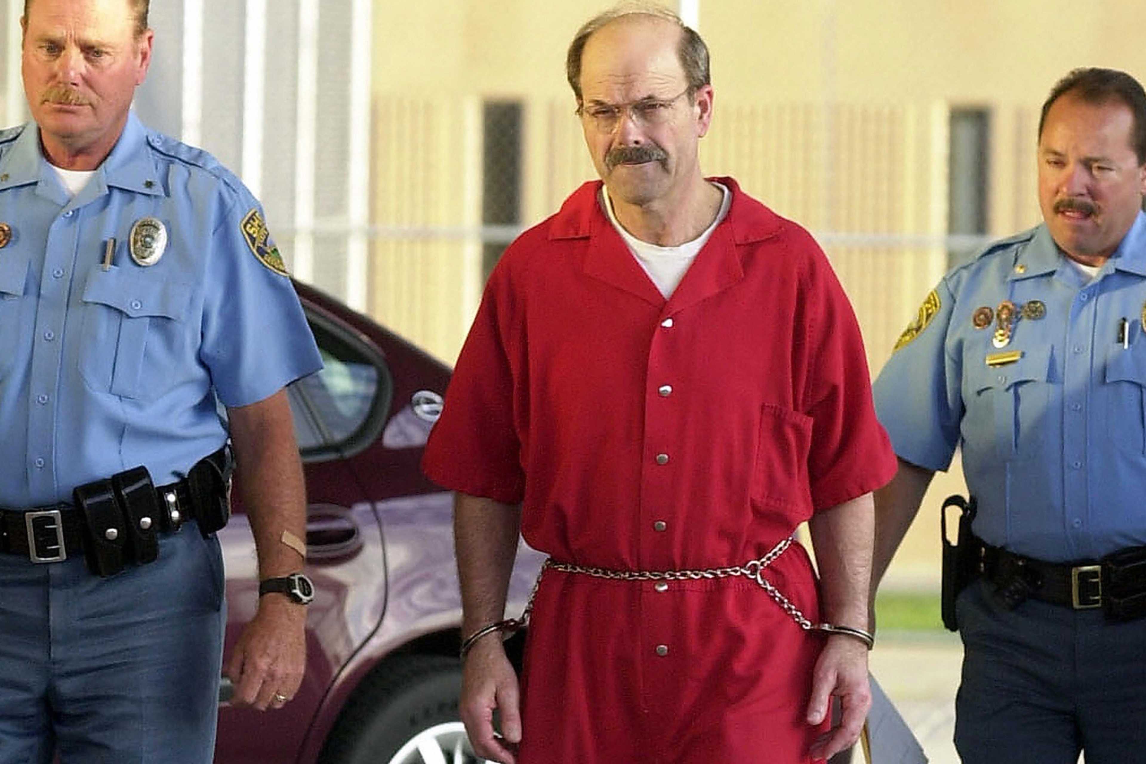 The image shows two police officers in uniform escorting a man wearing a red jacket, who appears to be in custody.