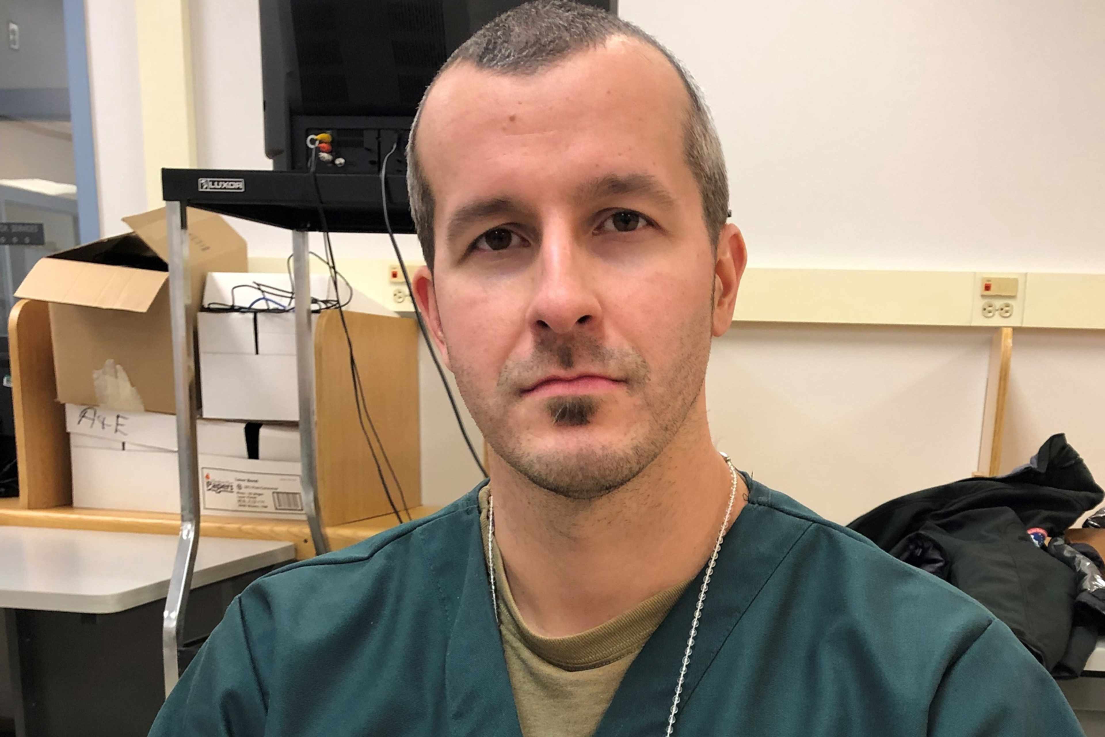 The image shows a man with a beard and short hair, wearing a green shirt, standing in what appears to be an office or workplace setting with various items in the background.