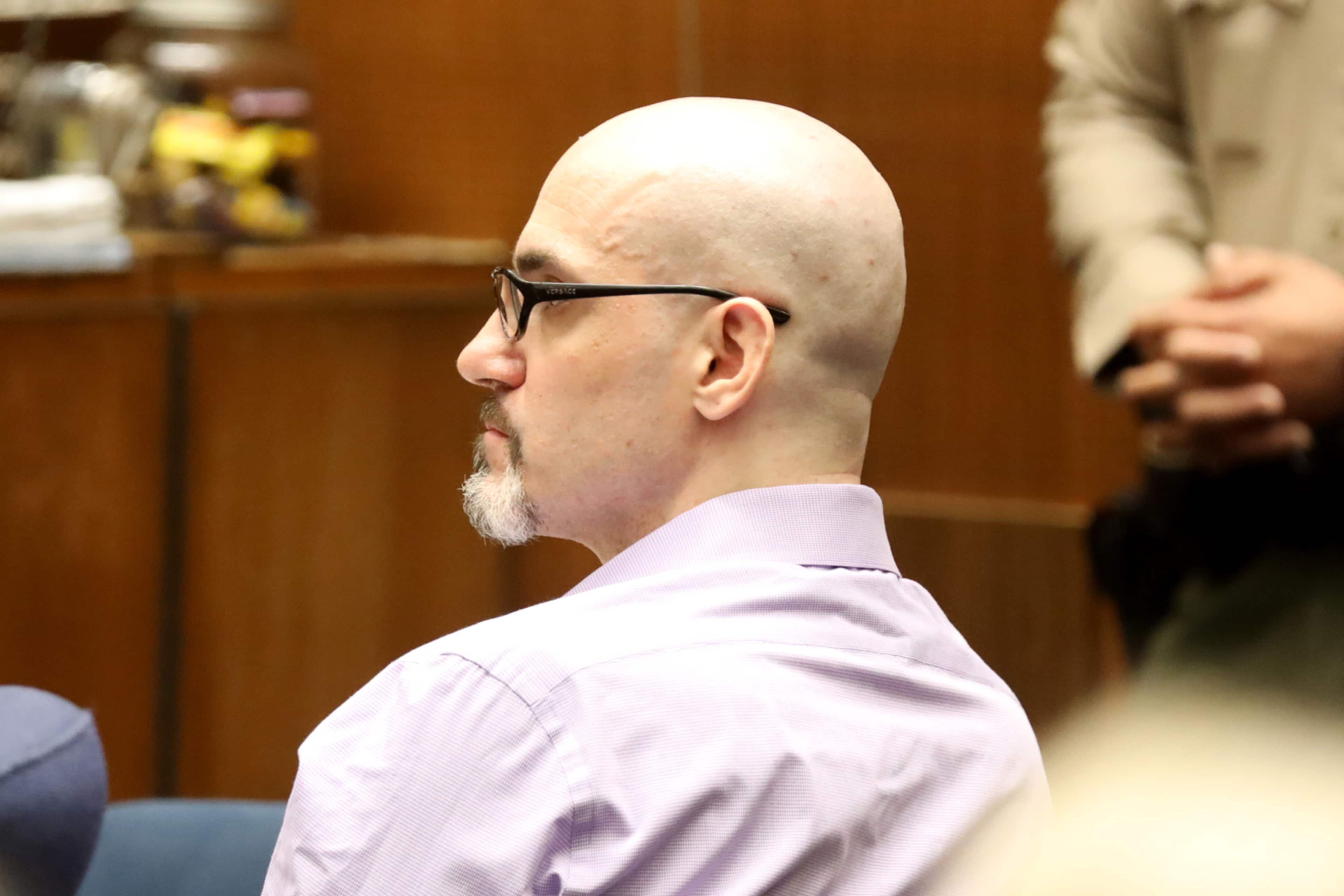 A bald man with glasses is seated in a courtroom, wearing a light-colored shirt and facing away from the camera.