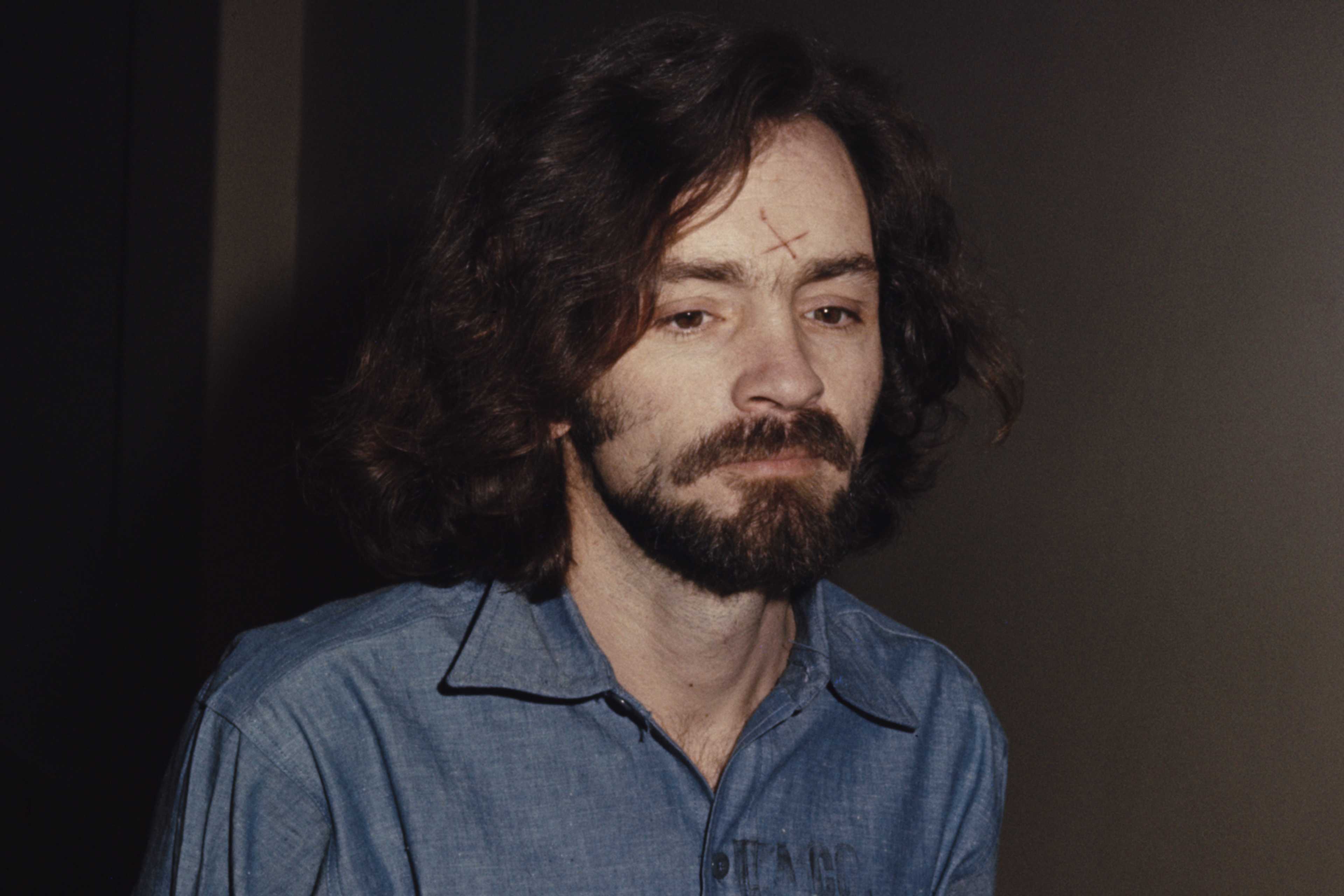 A man with long, dark hair and a beard is shown in a dimly lit setting, his expression appearing pensive or contemplative.