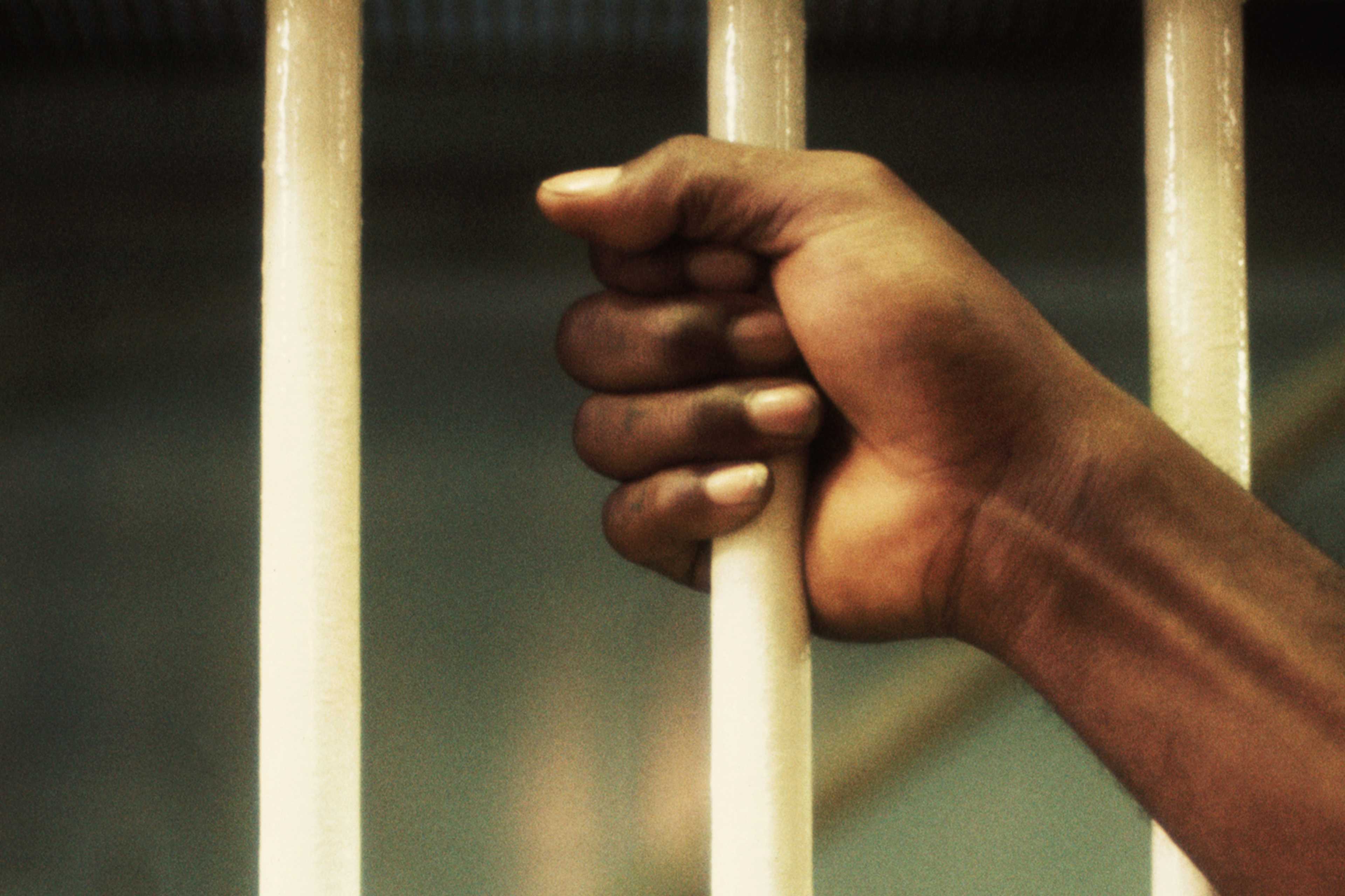 A hand gripping the metal bars of a prison cell, with a dark, blurred background suggesting a confined, gloomy environment.