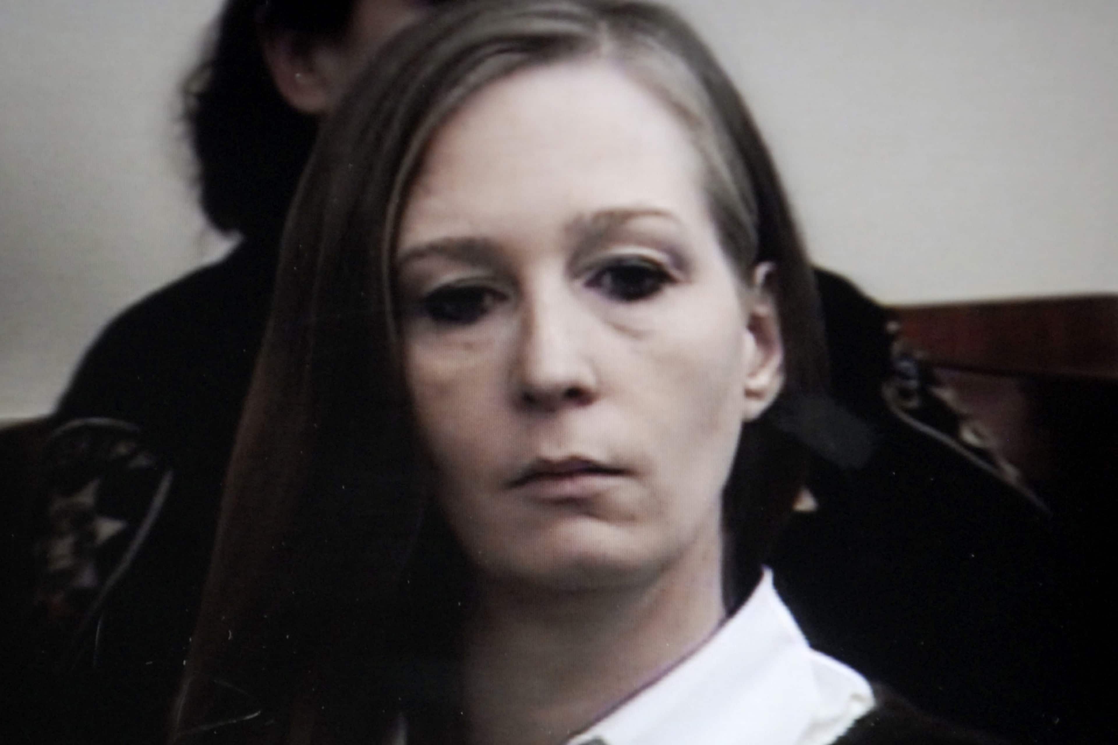 A woman with long, dark hair appears in the foreground, her expression somber and pensive, against a dimly lit background.