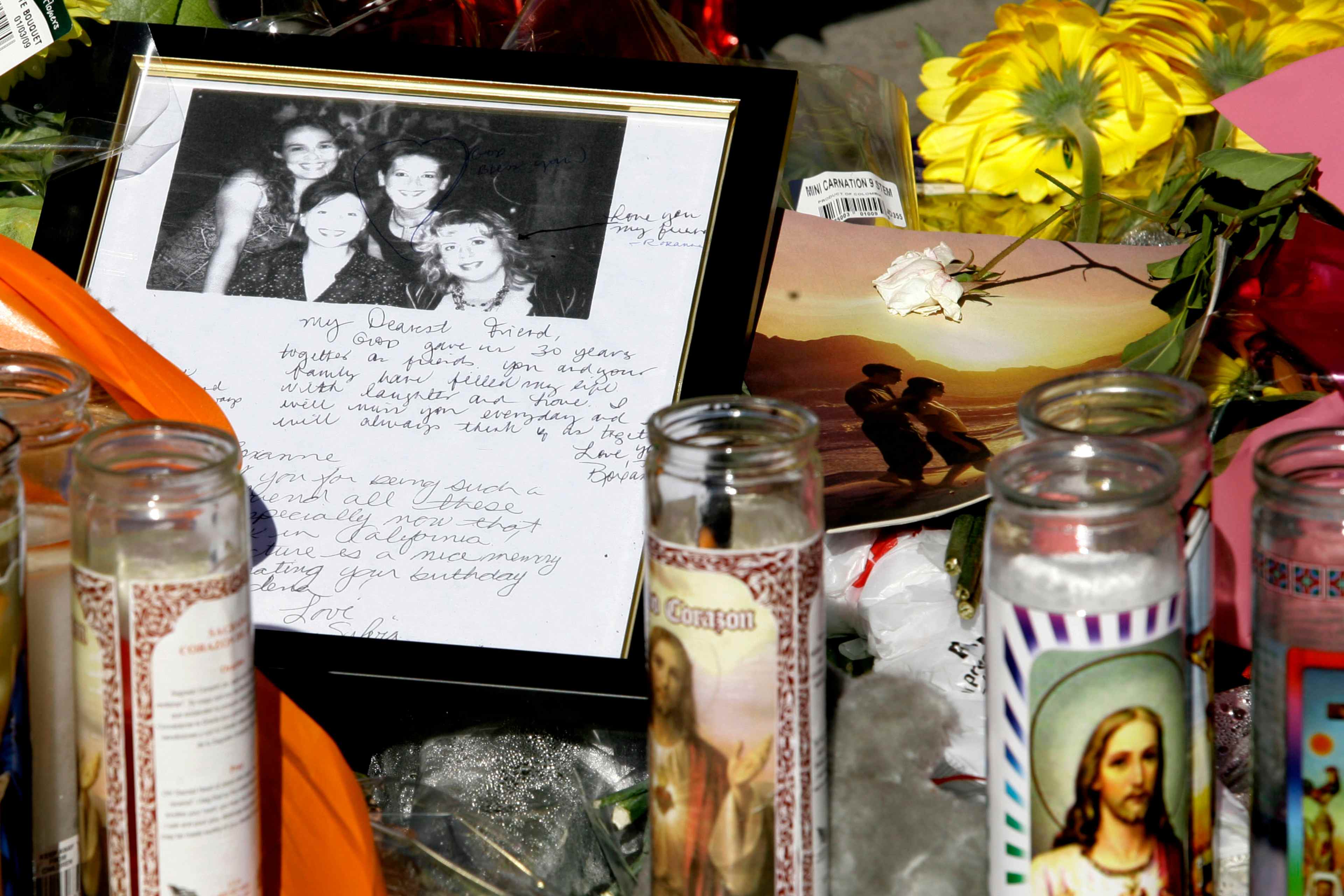 The image shows a framed photograph of several people surrounded by various memorial items, including candles, flowers, and handwritten notes, against a backdrop of colorful decorations.