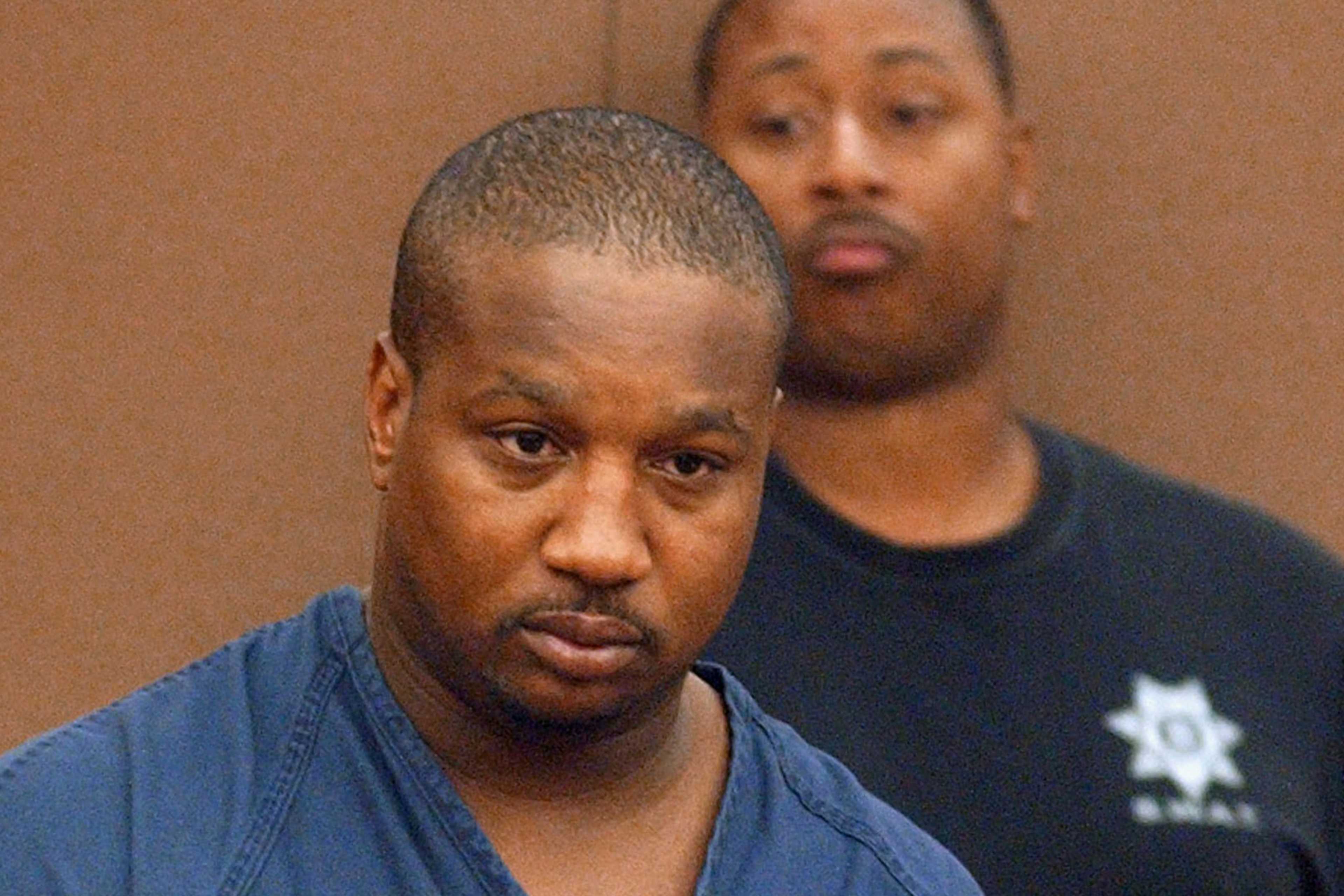 The image shows two African American men, one in the foreground with a serious expression and the other in the background with a concerned look.