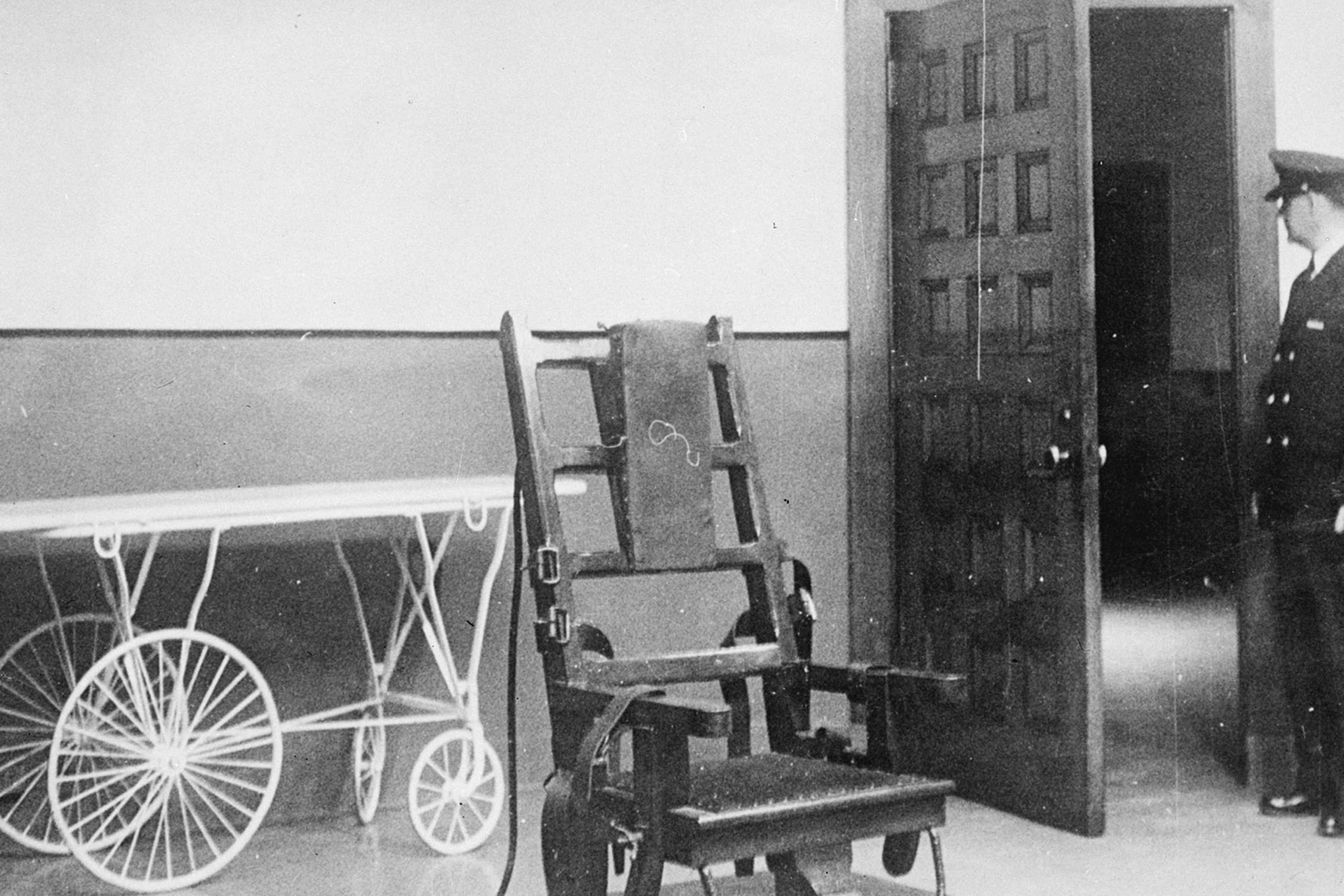 A black and white photograph showing a man standing in a doorway next to a bicycle and what appears to be a wooden chair or other furniture piece in the foreground.