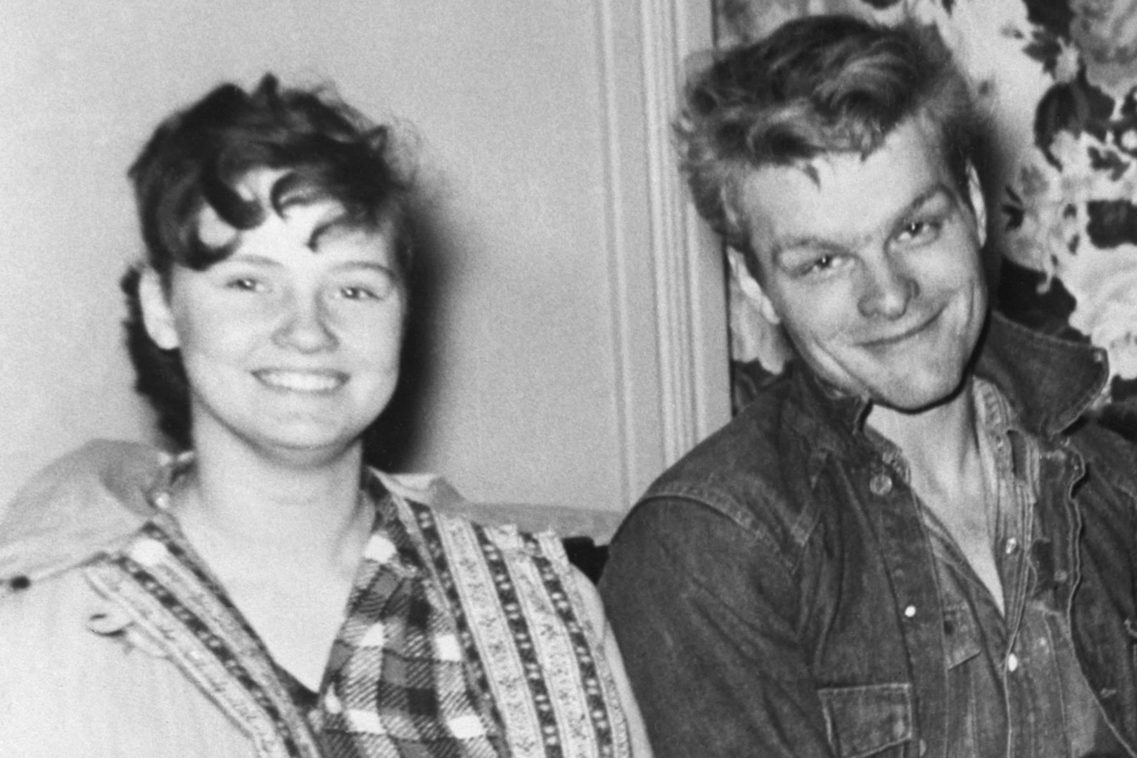 Two people, a man and a woman, are smiling and posing together in what appears to be a home setting with floral curtains visible in the background.