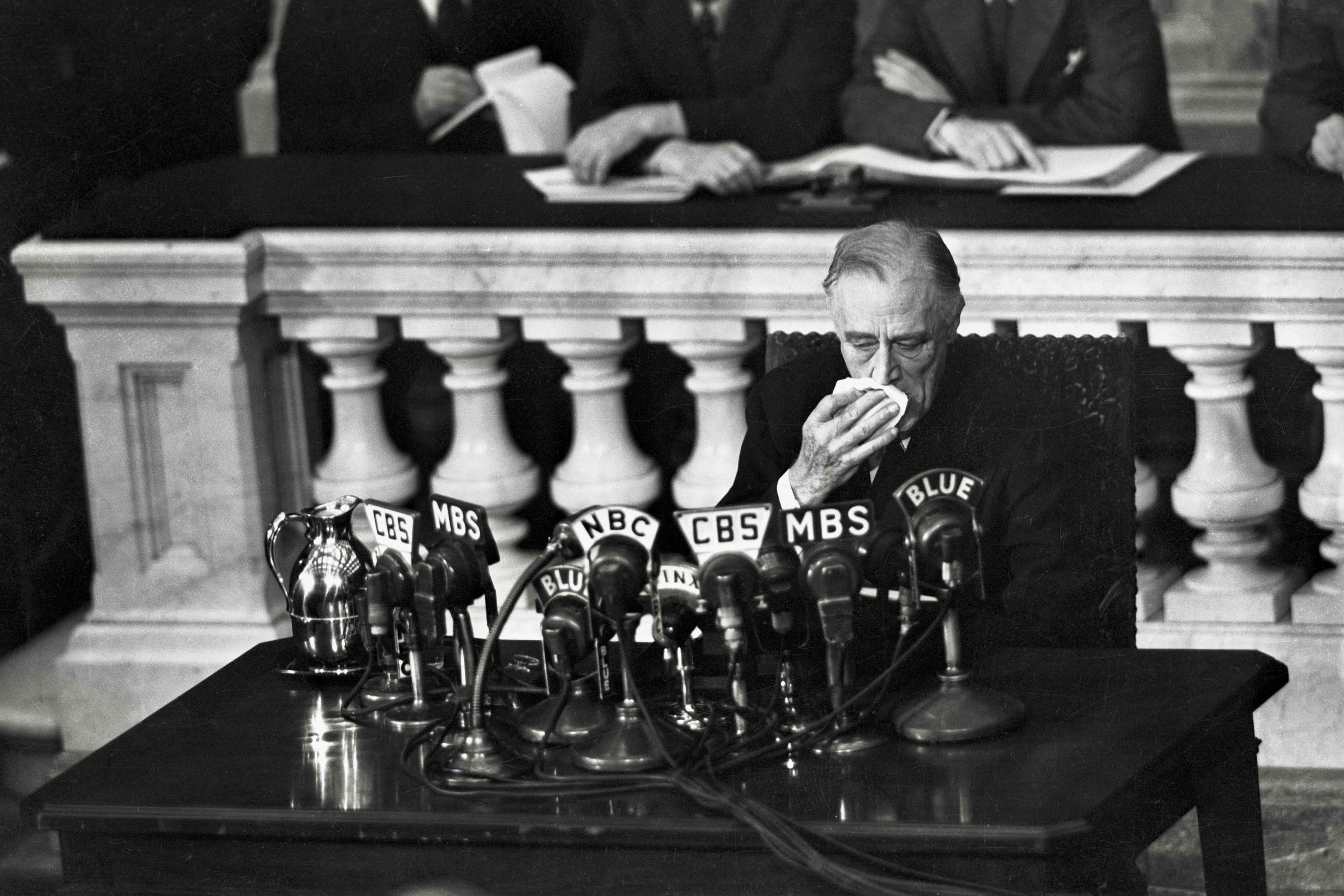 President Franklin D. Roosevelt is shown sitting at a table behind a crowded group of news microphones wiping his mouth with a handkerchief during his reporting to the House of Representatives on the 'Big Three' conference in Yalta.