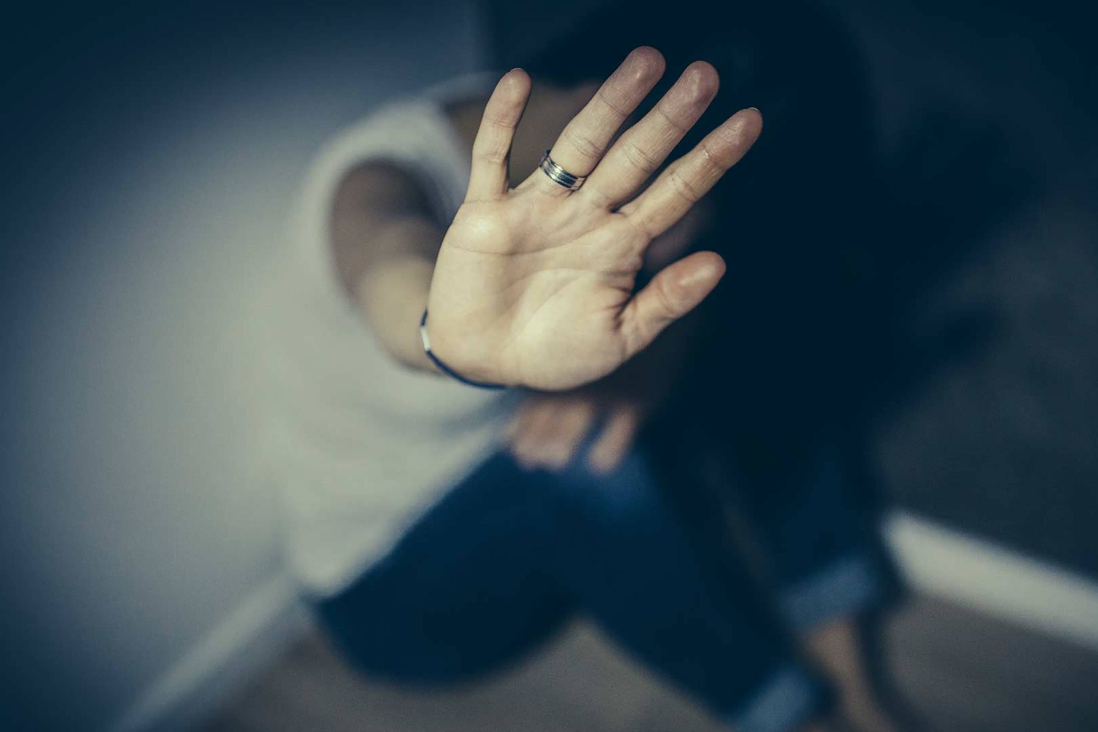 A hand is raised in a gesture of defense or protection against a dark, blurred background.