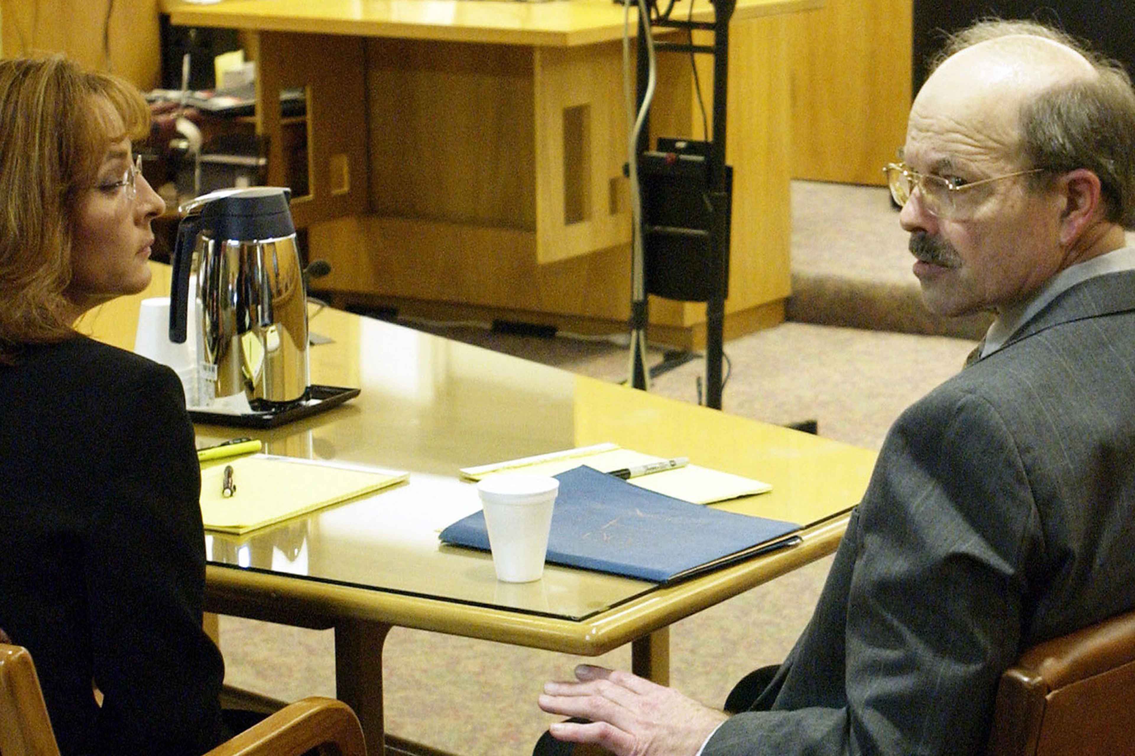 Two individuals, a woman and a man, are engaged in a conversation at a desk in what appears to be an office or professional setting.