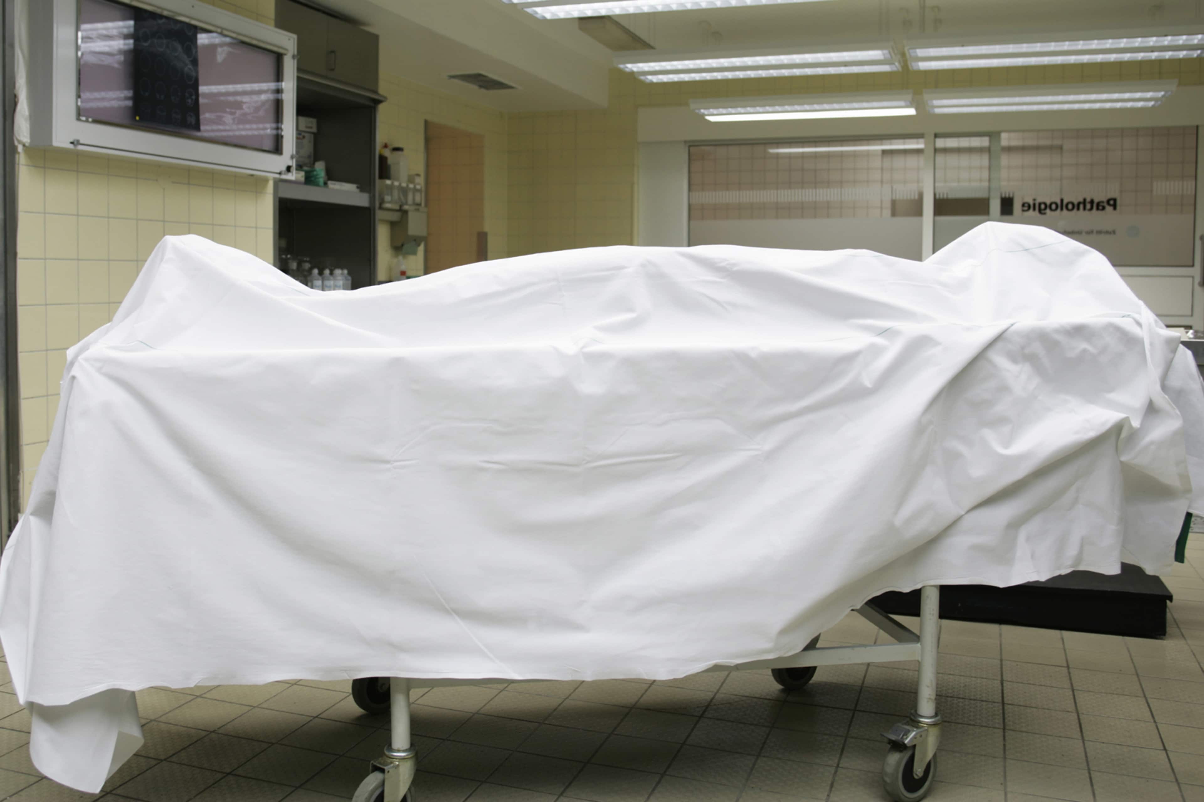A white sheet covers a hospital bed or gurney in a tiled, institutional-looking room with fluorescent lighting.