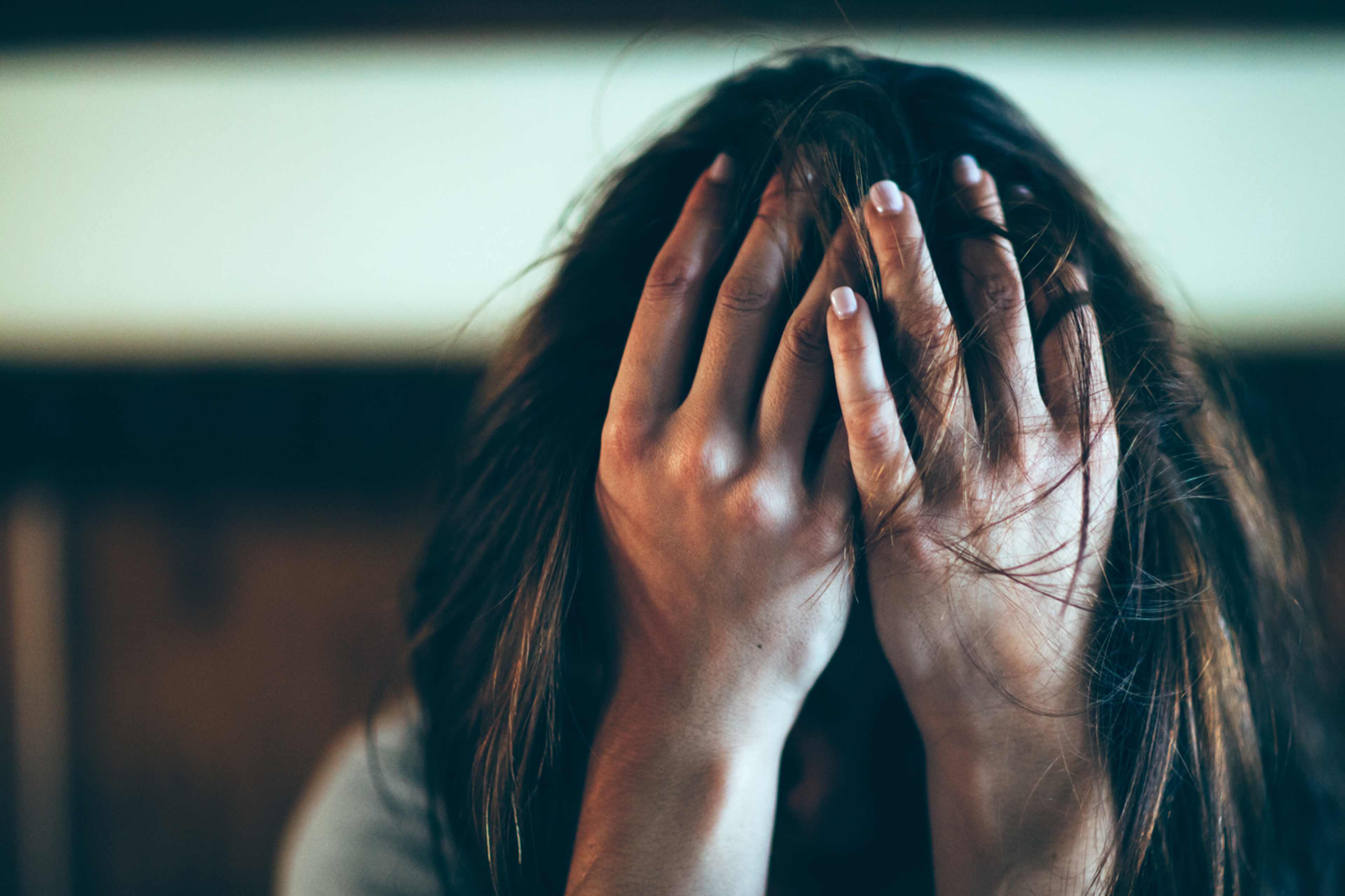 The image shows a person's hands covering their face, with long dark hair visible between the fingers. The background appears to be a dimly lit room or space.