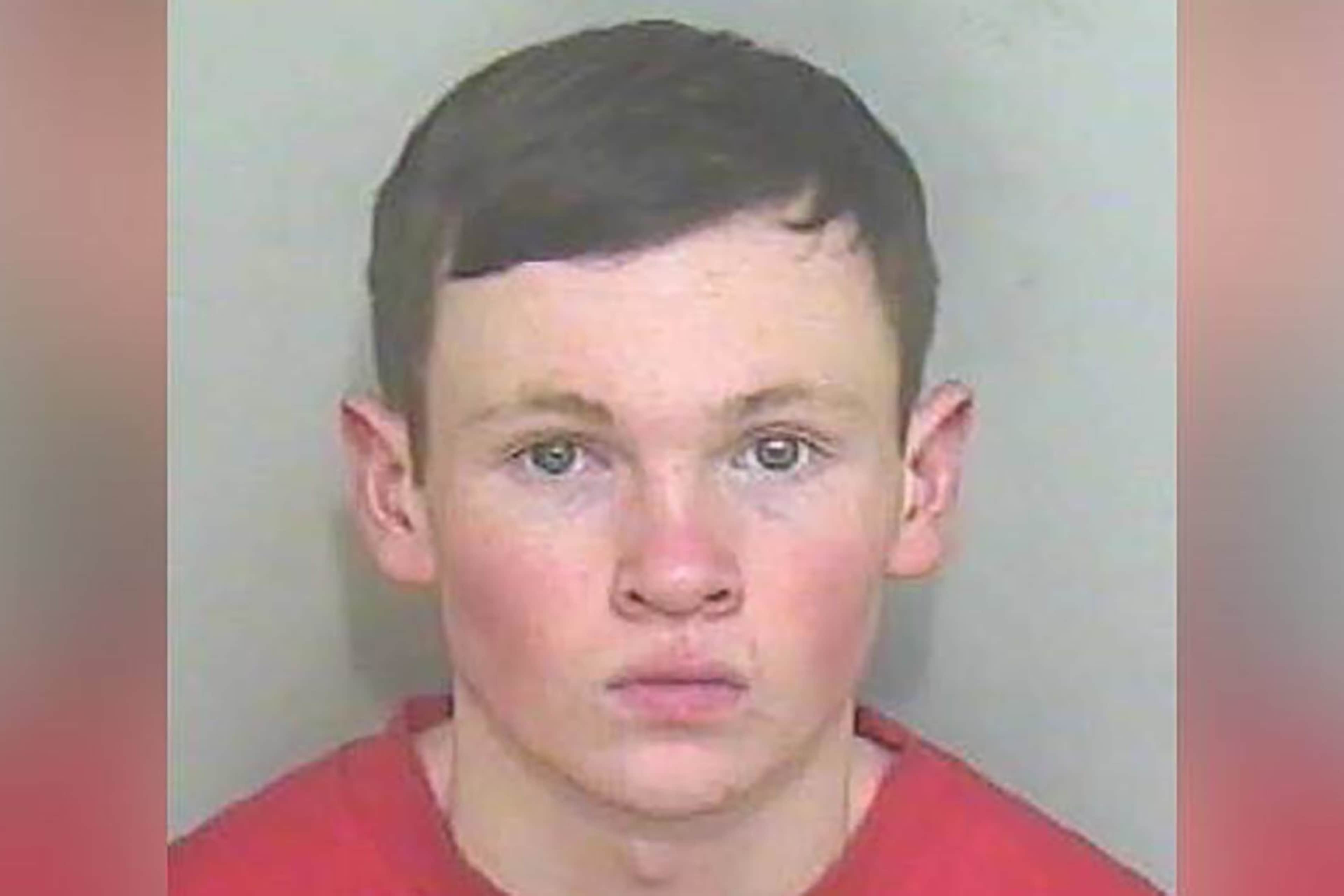 The image shows a close-up portrait of a young man with short dark hair and a serious expression, set against a plain pink background.