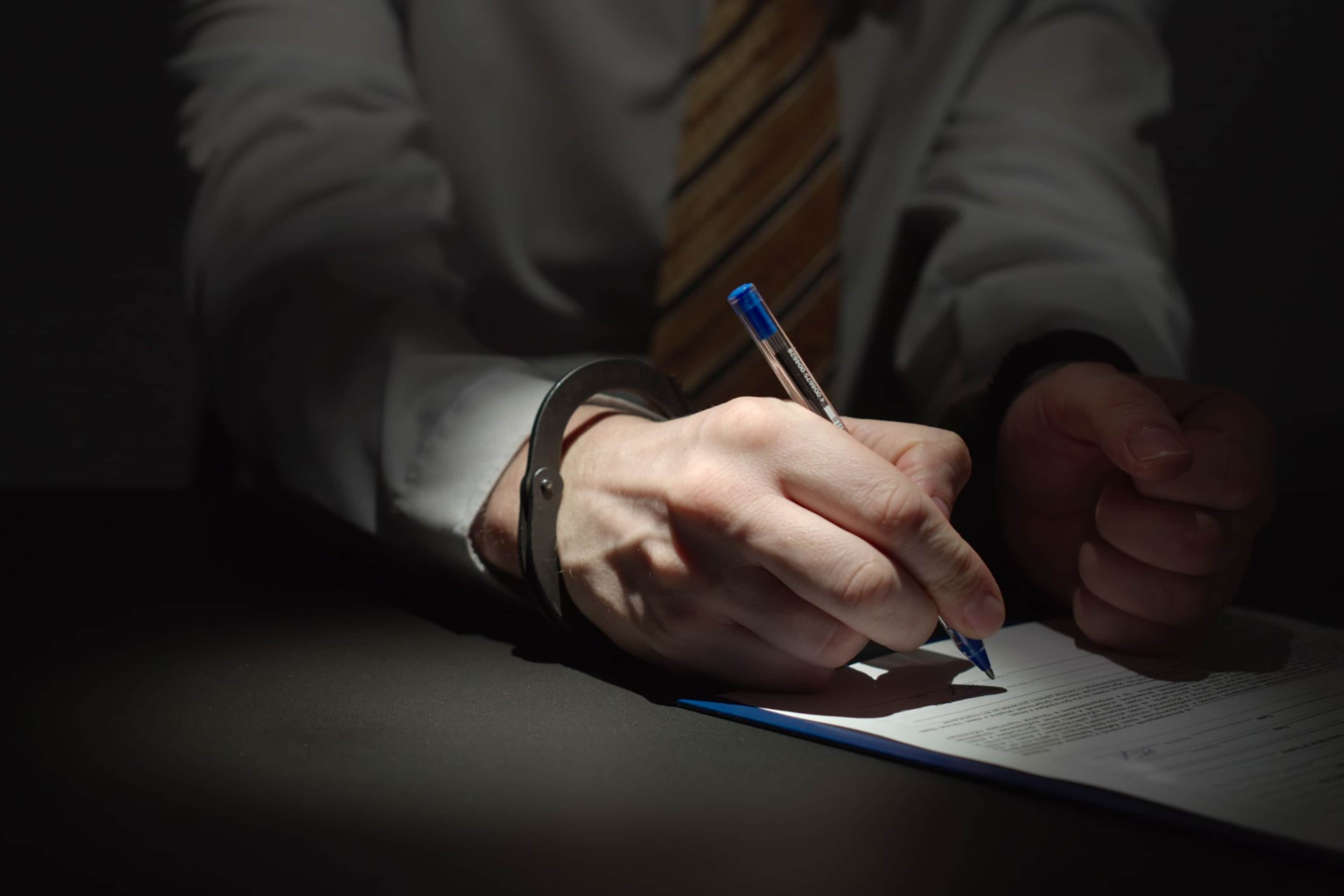 A person's hands holding a pencil and writing on a document in a dimly lit environment.
