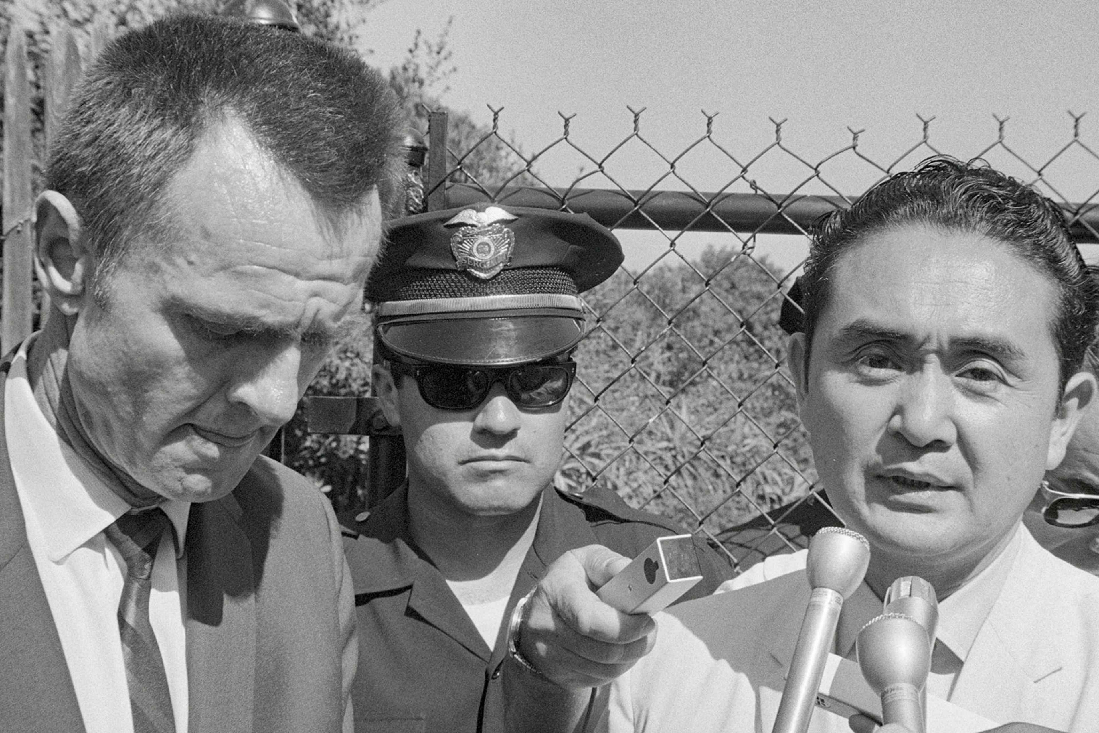 The image depicts three men, one wearing a police uniform, standing in front of a wire fence with barbed wire at the top.