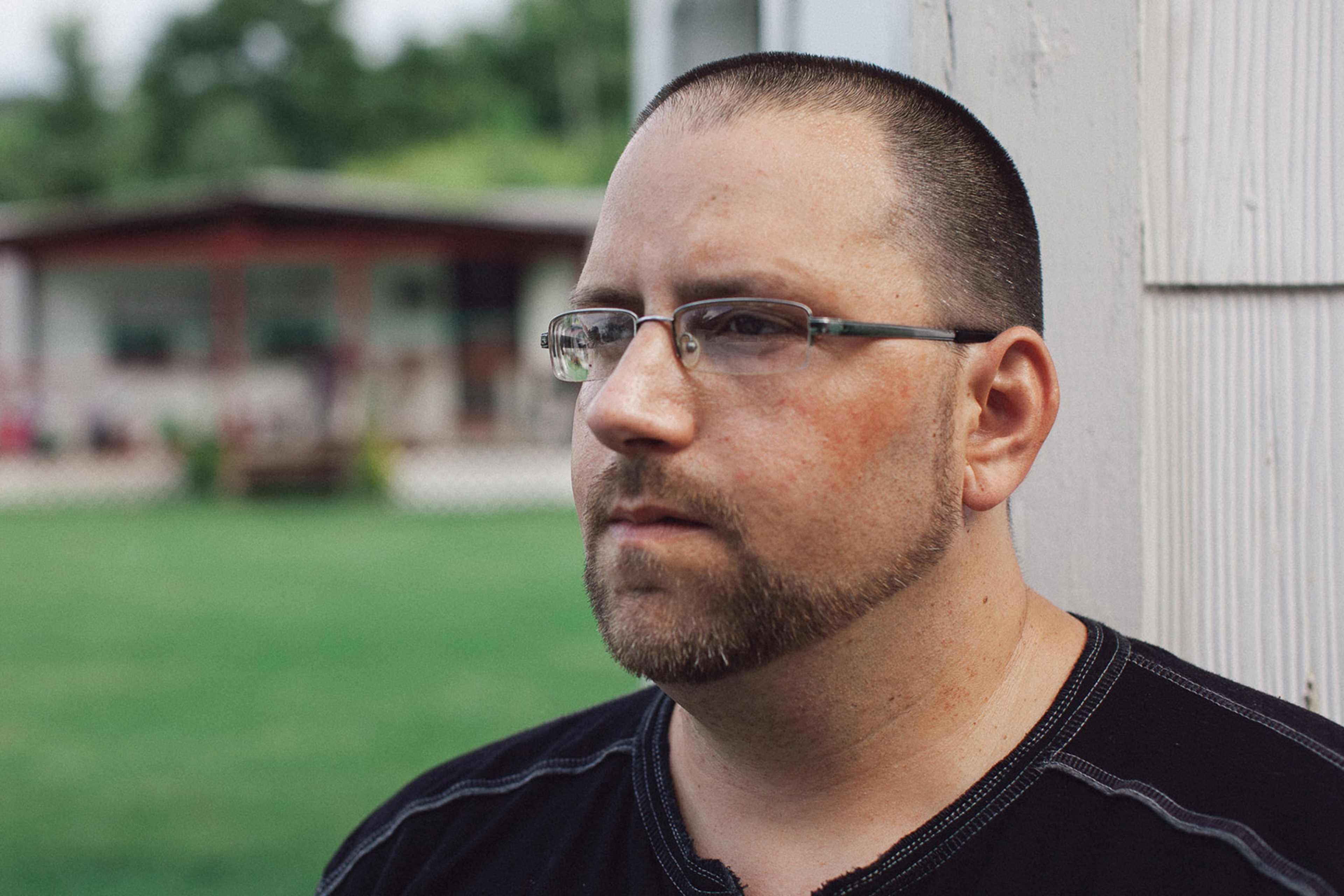 A man with a beard and glasses is standing in front of a building with a grassy area in the background.