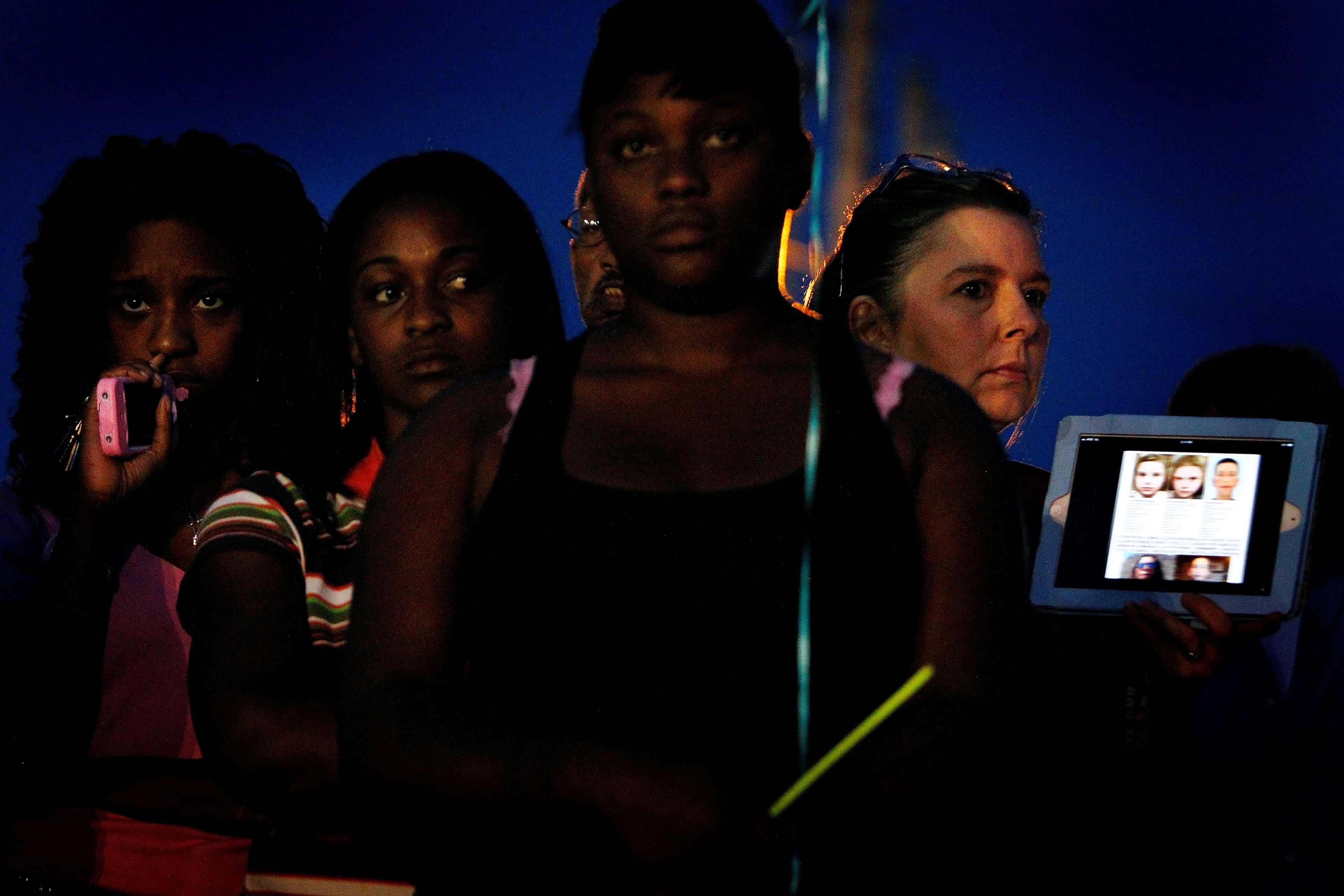 The image shows a group of people, including a woman holding a digital device displaying several faces, against a dark, nighttime background.
