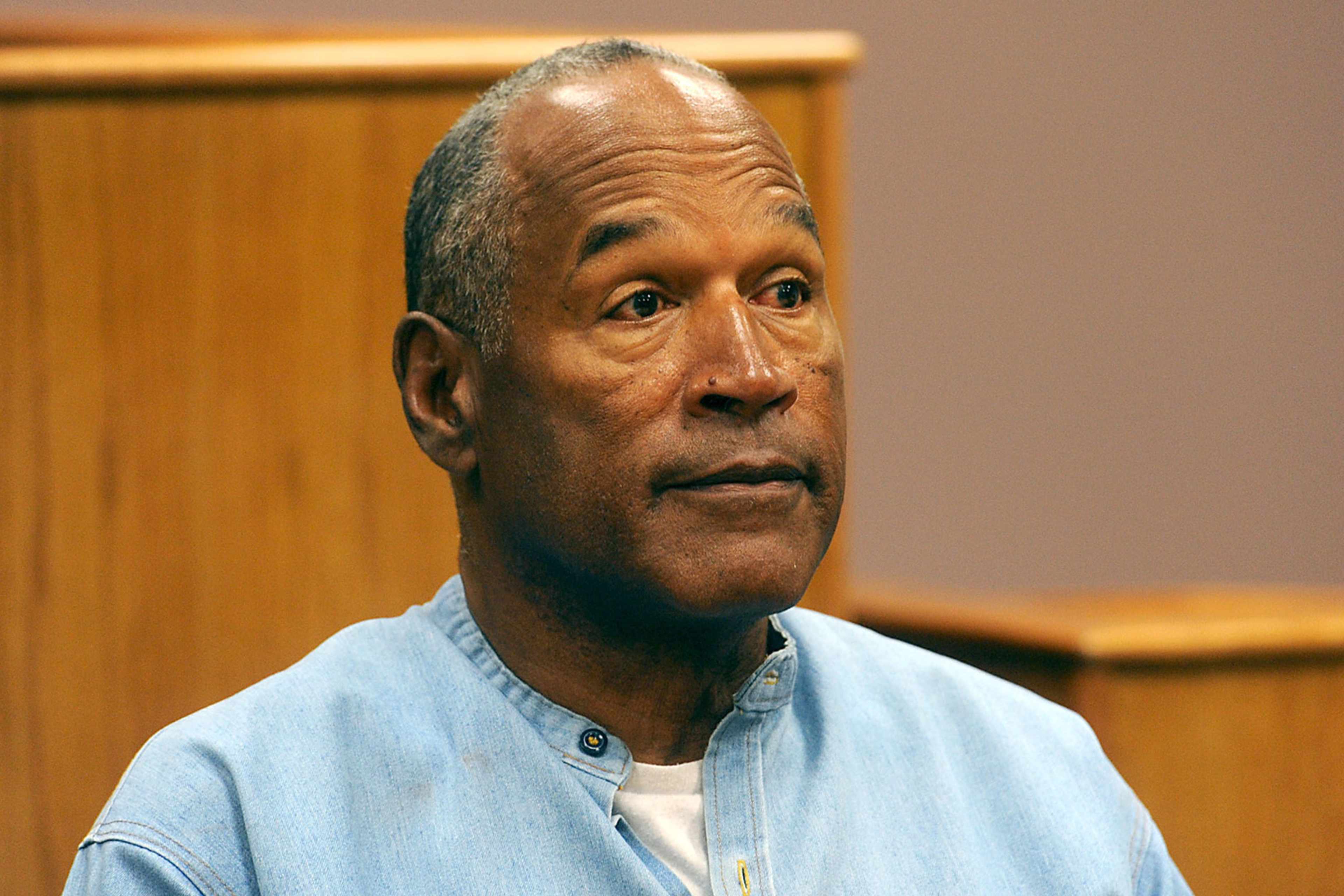 The image shows an elderly African American man with a serious expression, wearing a light blue shirt, seated in what appears to be a courtroom or similar formal setting.