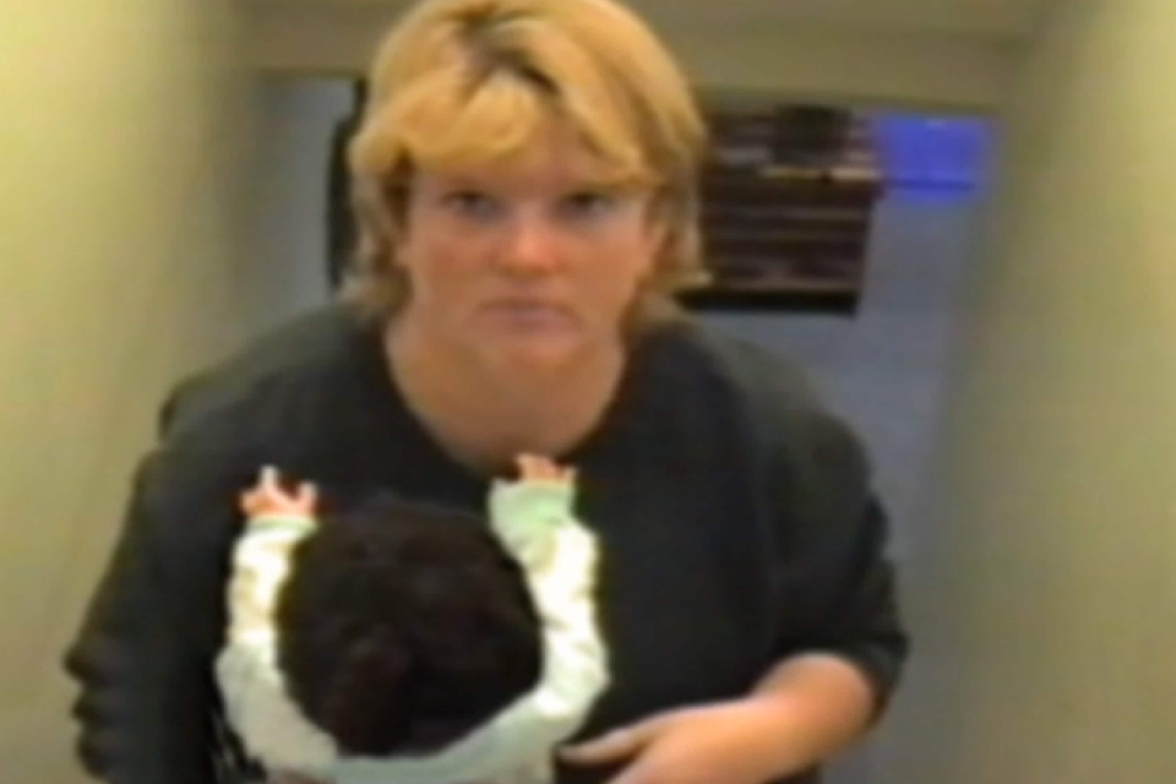 The image shows a woman with blonde hair wearing a black jacket, standing in what appears to be a hallway or indoor setting.