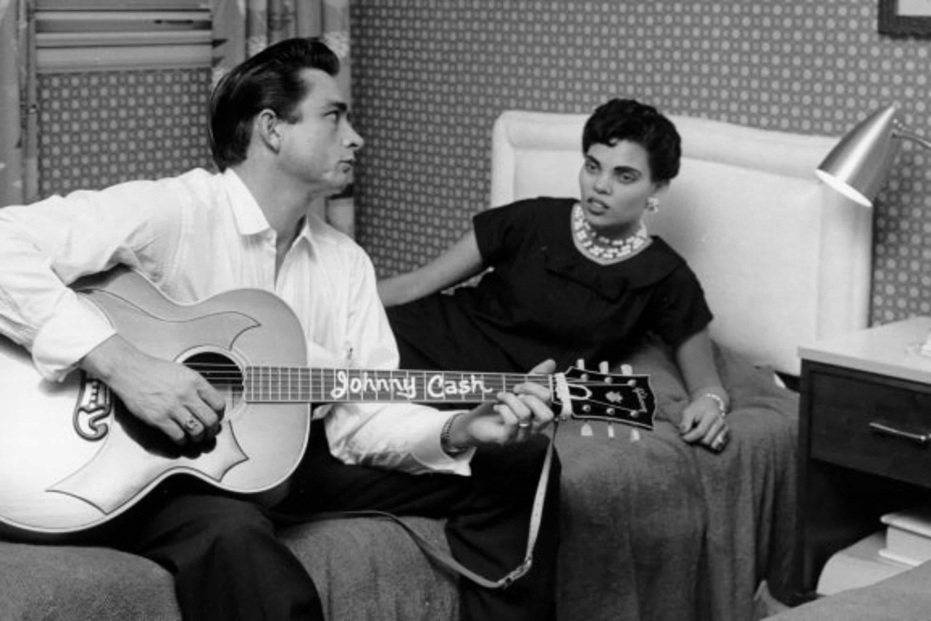 Country singer/songwriter Johnny Cash sits on a bed playing acoustic guitar to his first wife Vivian Liberto