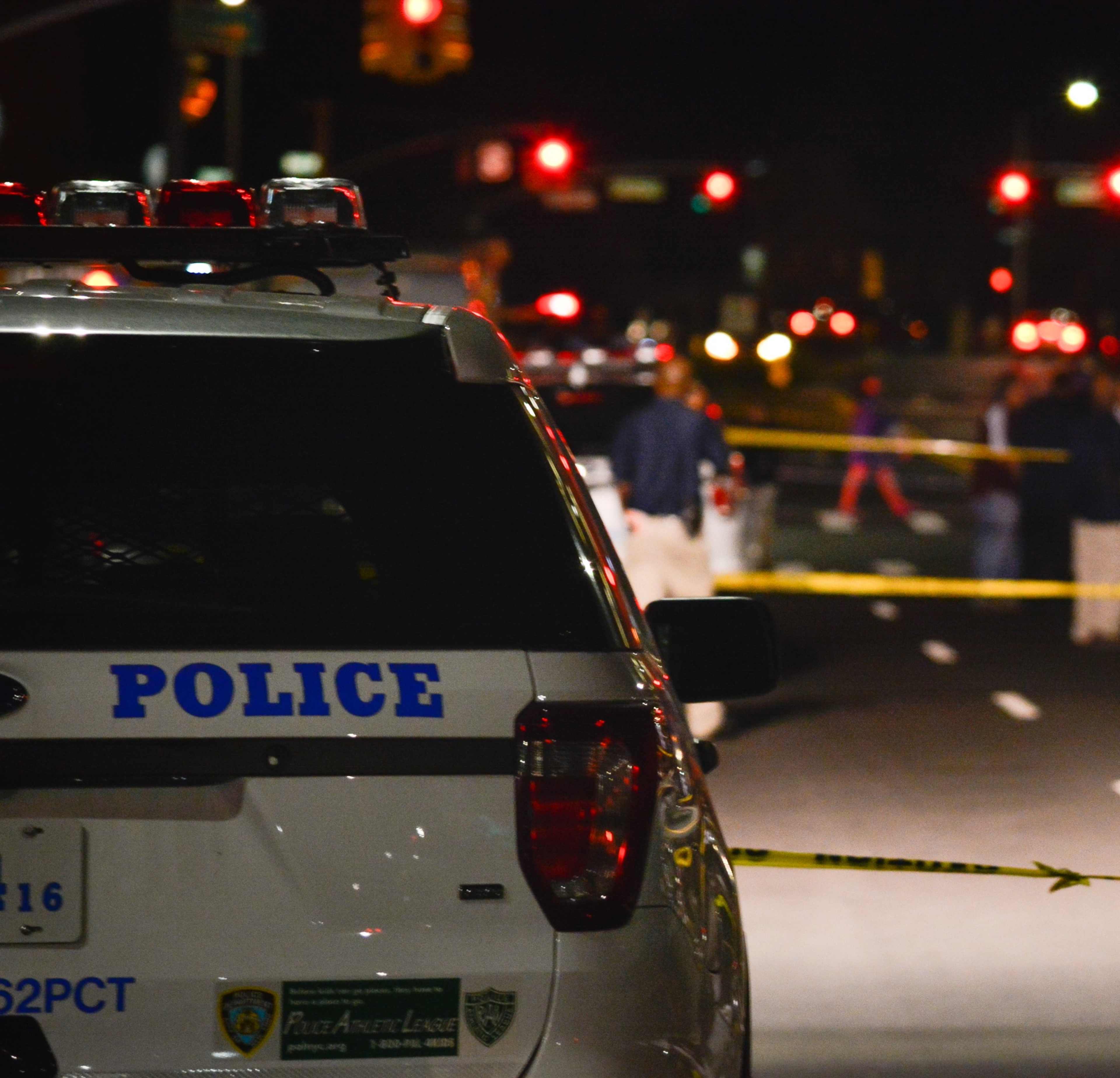 A police vehicle is parked on a dimly lit street, with emergency lights flashing and police officers visible in the background, suggesting an ongoing investigation or incident.