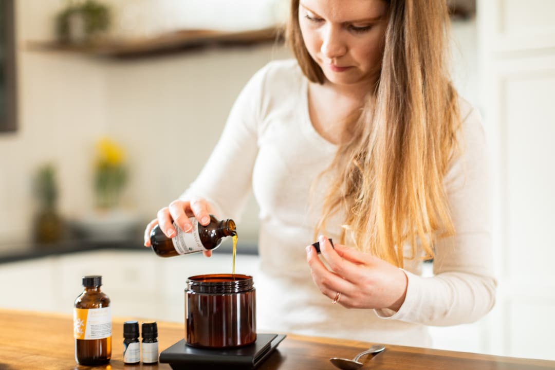 Pouring golden jojoba oil from an amber bottle into a jar on a kitchen scale