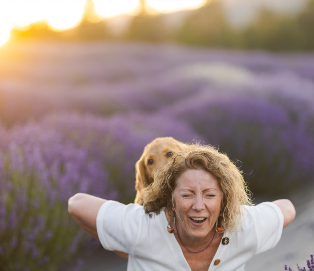 Woman laughing with her arms spread wide in a blooming lavender field at sunset, a golden retriever at her shoulder