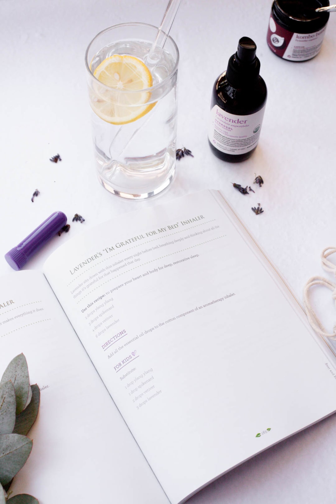 A purple aromatherapy inhaler beside an open recipe book, a lavender hydrosol bottle, and scattered dried lavender buds