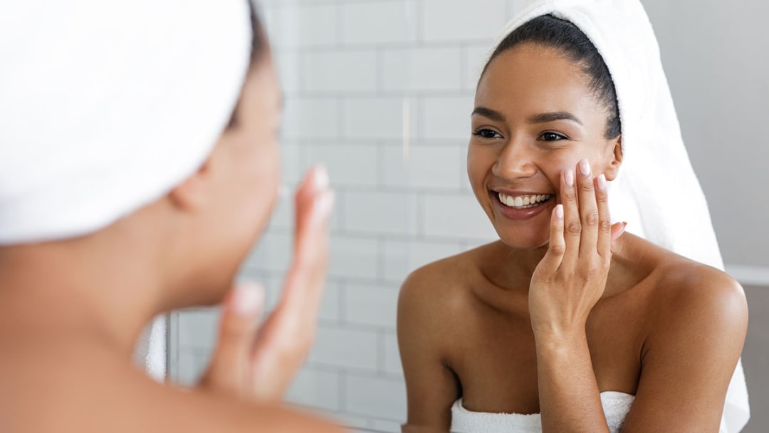 A woman with her hair wrapped in a towel smiling into a bathroom mirror as she presses oil into her cheek