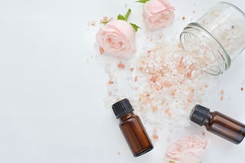 Pink rock salt spilling from a tipped glass jar beside two amber essential oil bottles and pale roses